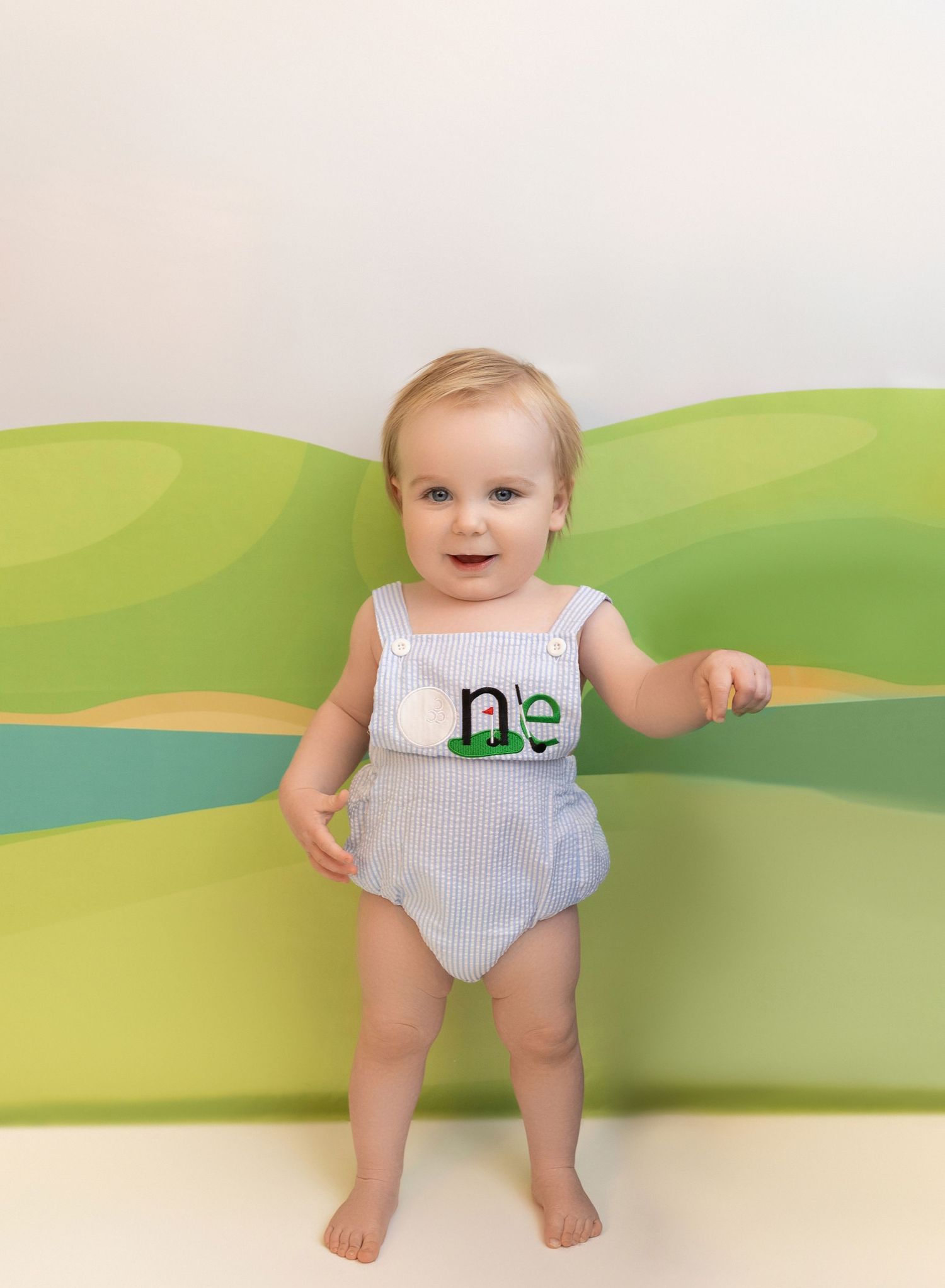 A baby in seersucker romper sits against a whimsical green wall mural depicting rolling hills and a river.