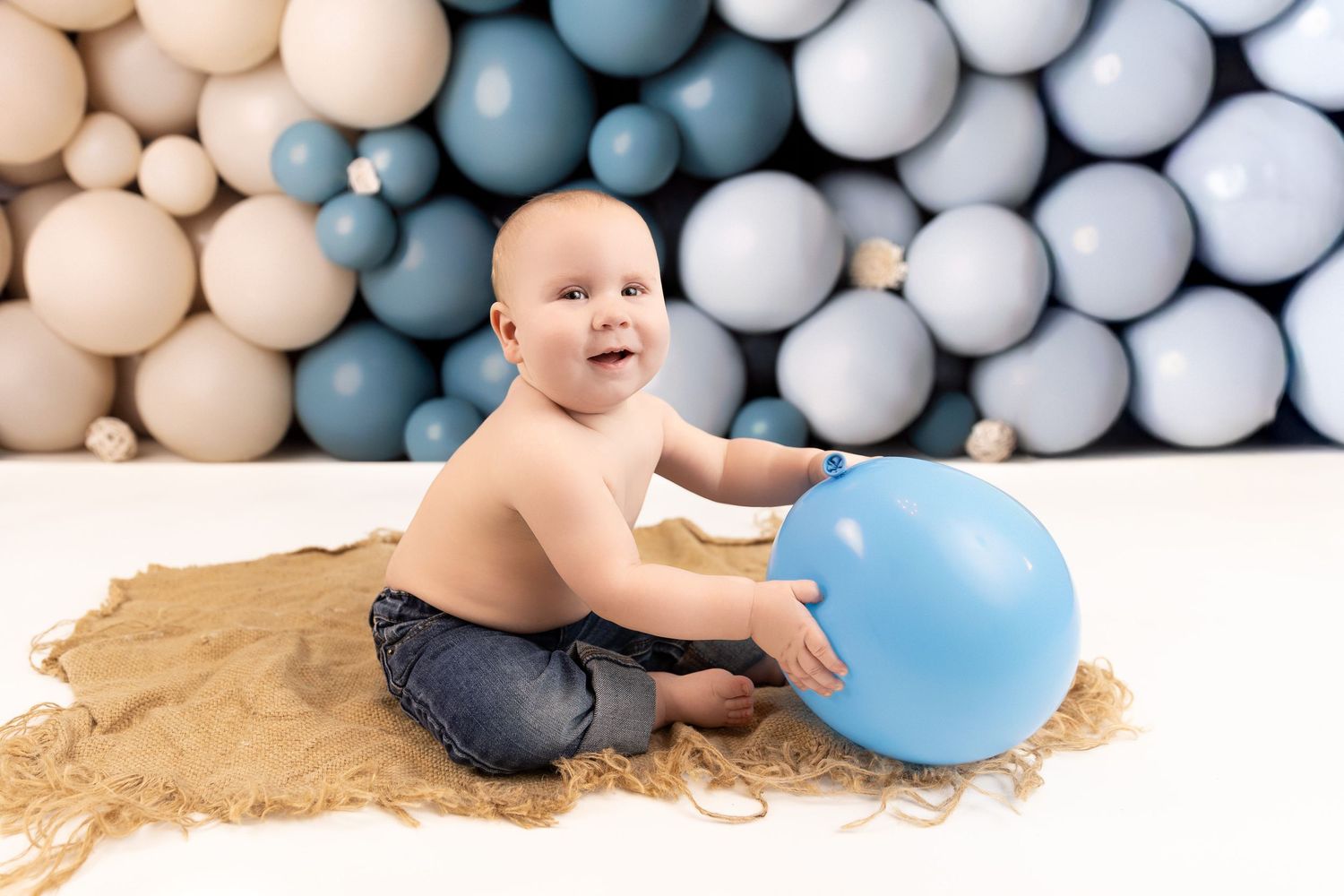 Baby sits in a ball pit with soft blue and beige balls while playing with a large balloon.