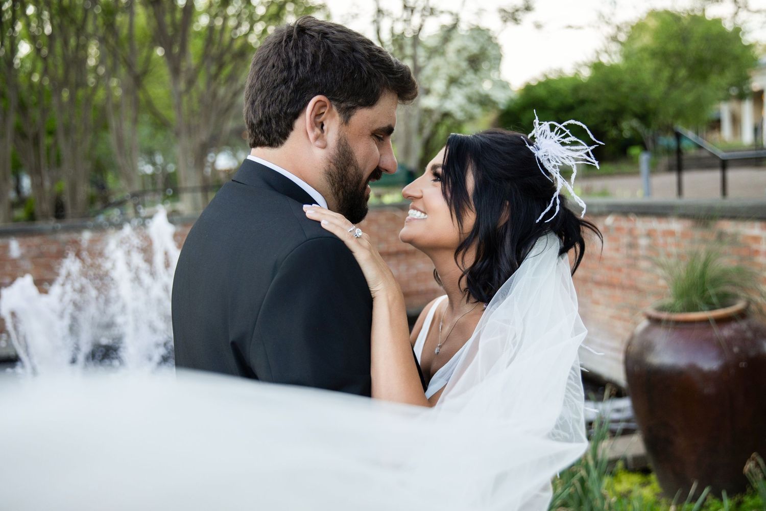 Bride and groom share a romantic moment near a fountain, with her veil flowing in a lush garden wedding setting.