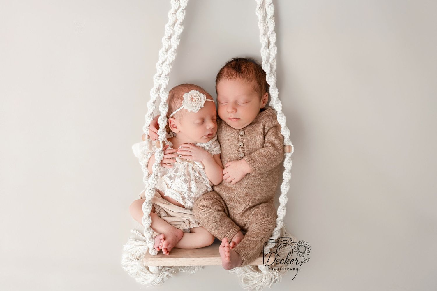 Two sleeping babies wearing beige outfits rest peacefully together on a white rope swing against a neutral background.