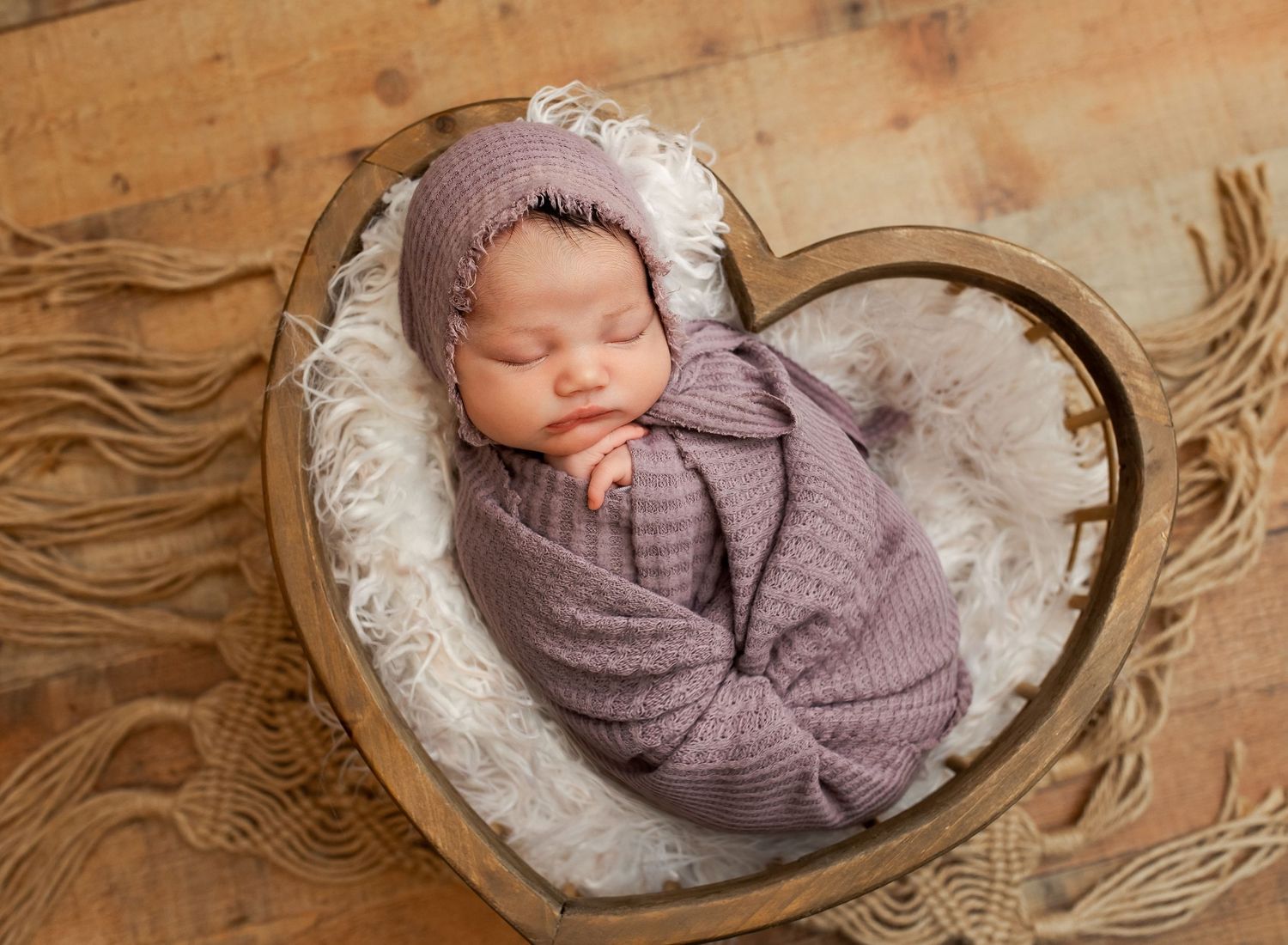 Sleeping newborn baby wrapped in purple swaddle with matching bonnet resting in heart-shaped wooden bowl with white fur.