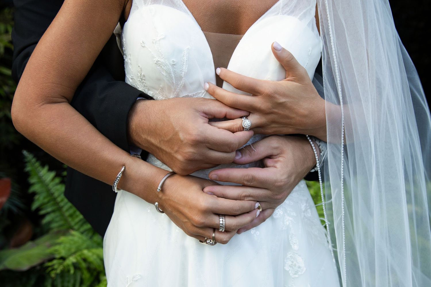 Newlywed couple holding hands showing wedding rings, bride in white gown with veil, groom in dark suit.