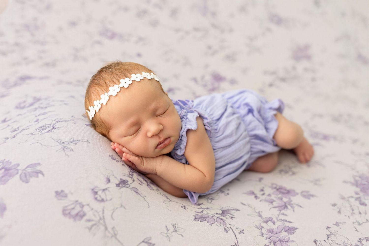 Sleeping newborn baby in lavender romper with floral headband on purple floral blanket.