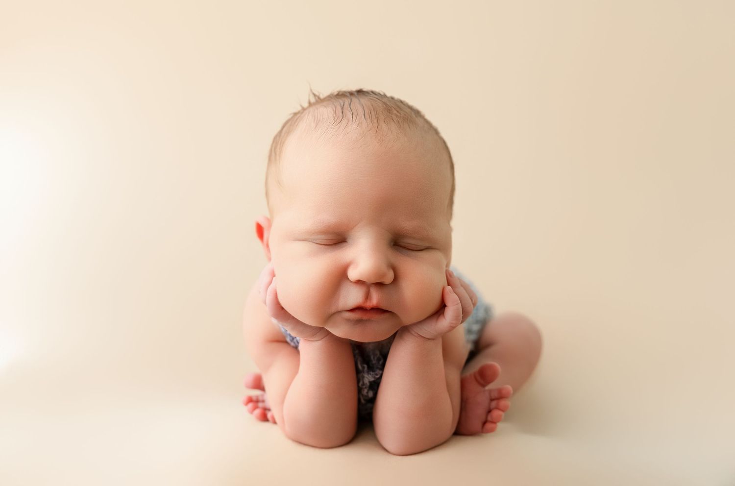 Sleeping newborn baby posed with hands under chin on cream background.