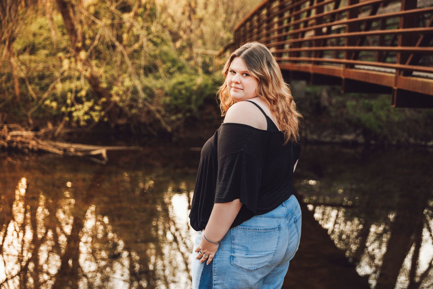 Woman in black off-shoulder top and jeans posing by a wooden bridge over a reflective creek at golden hour.