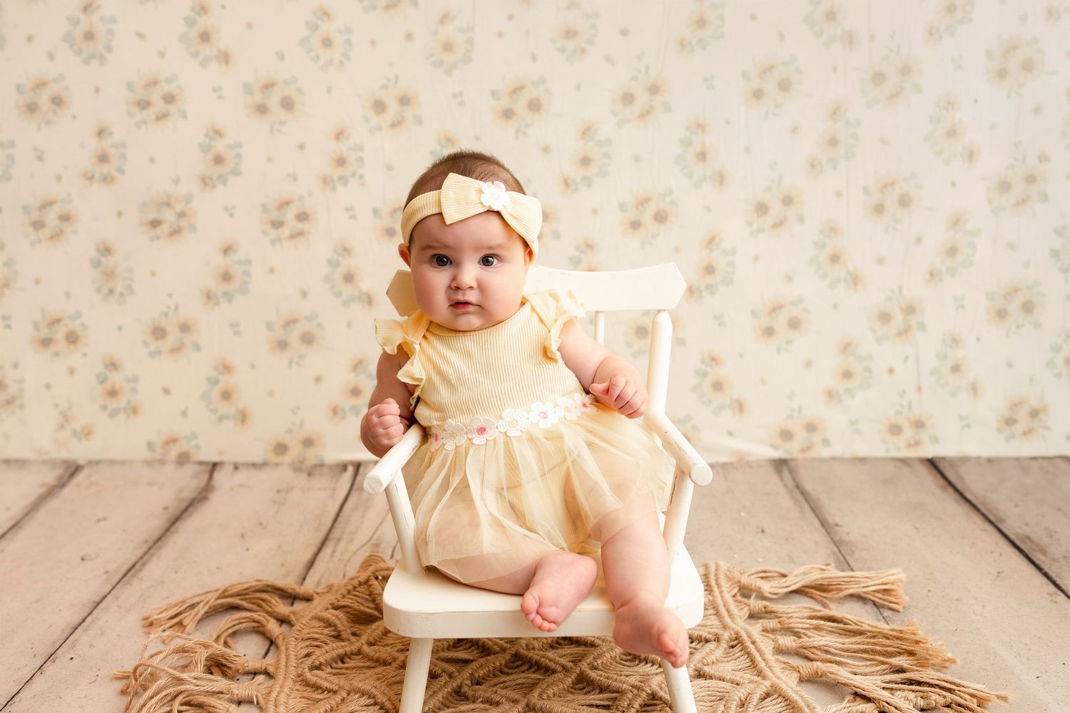 Baby girl in a yellow tulle dress and bow headband sits on a small white wooden chair against a floral backdrop on a rustic wood floor.