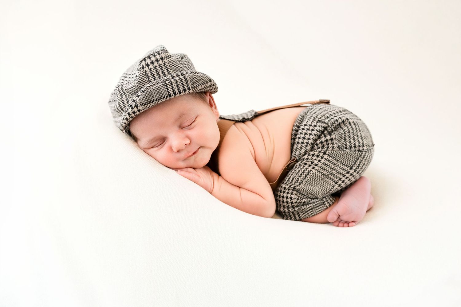 A sleeping newborn wearing a gray plaid newsboy cap rests peacefully on a white backdrop.