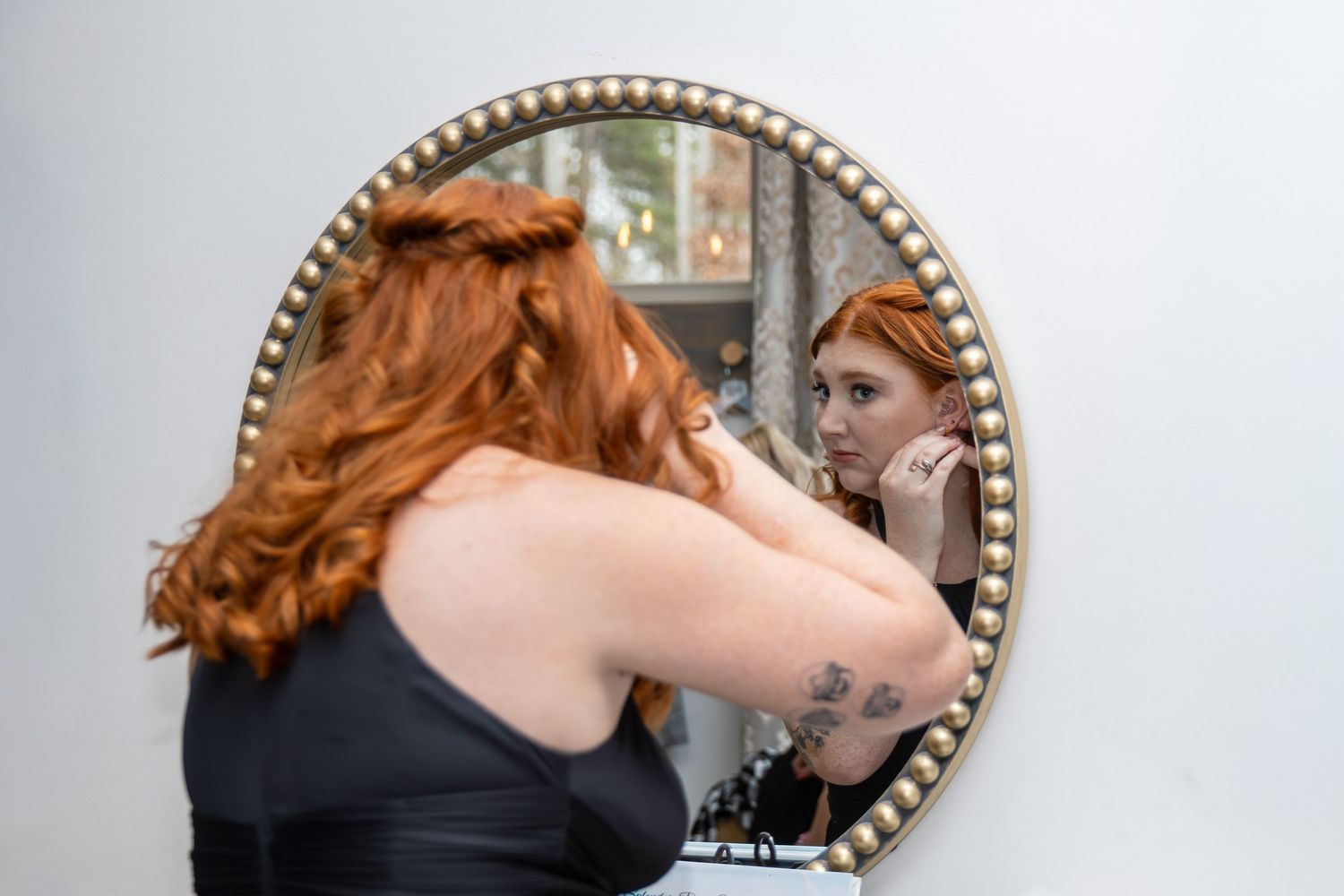 Red-haired woman with tattoos adjusting earring while looking at her reflection in a decorative round mirror.