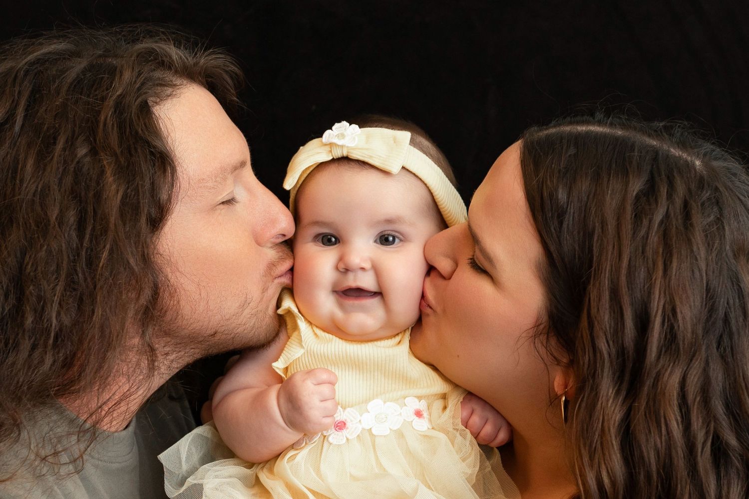 Two parents kissing their smiling baby girl wearing a yellow dress and white headband bow.
