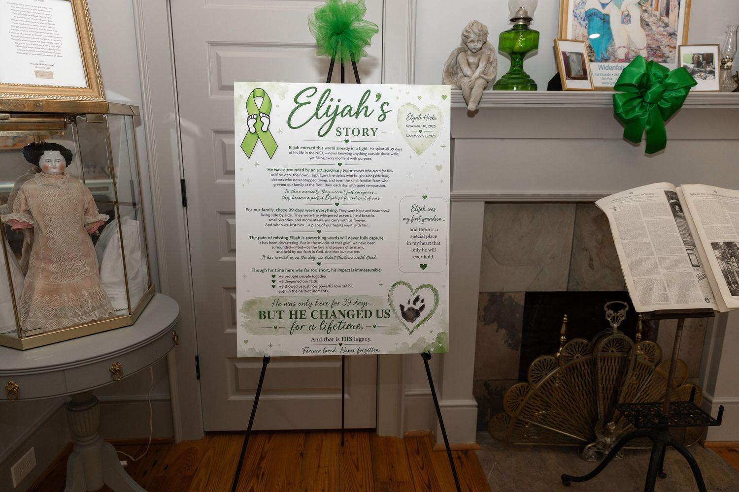 A memorial display board titled Elijah's Story on an easel, featuring green ribbon decorations in a decorated room.
