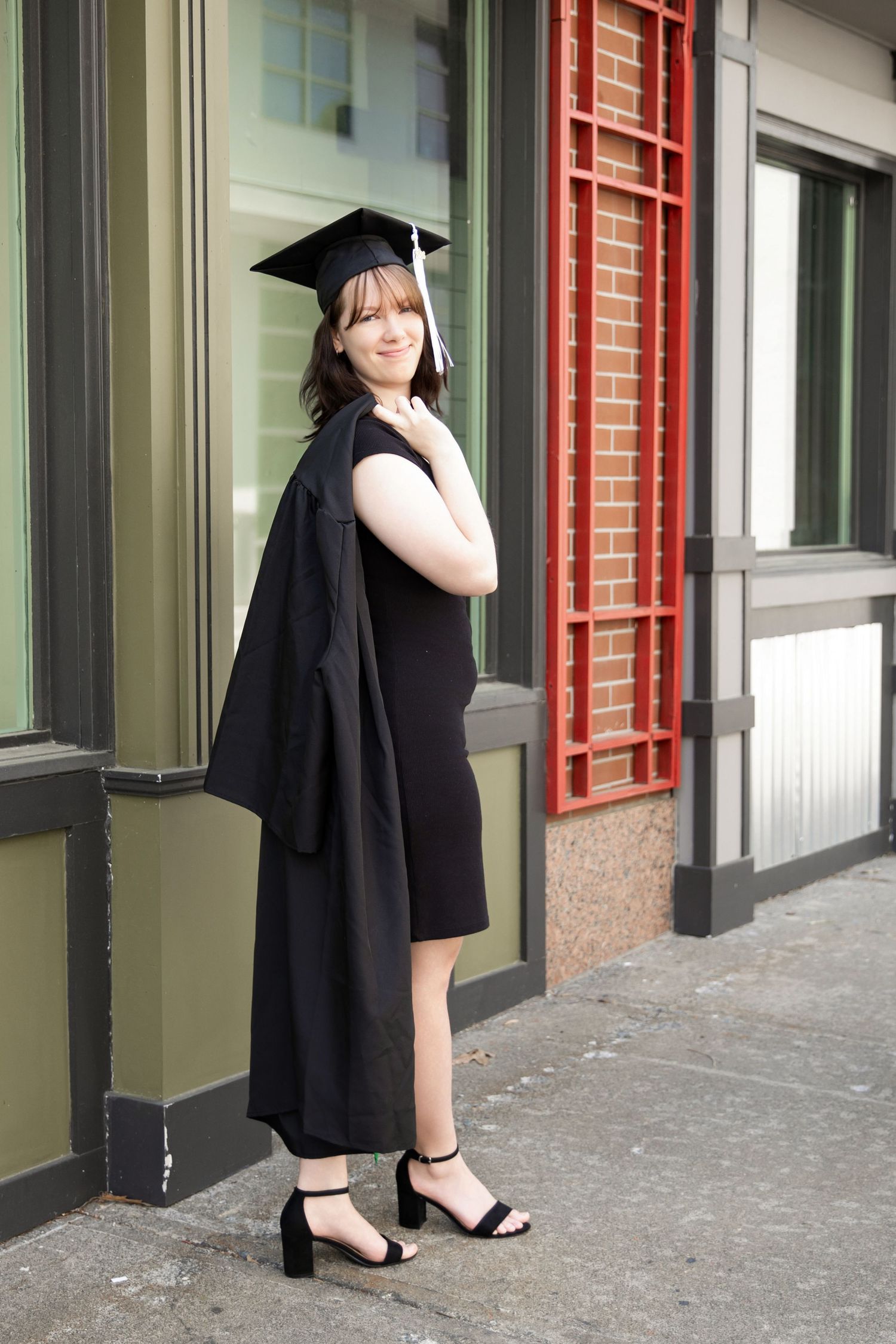 Young woman in black graduation cap and gown holding robe over shoulder, smiling outdoors near building windows.