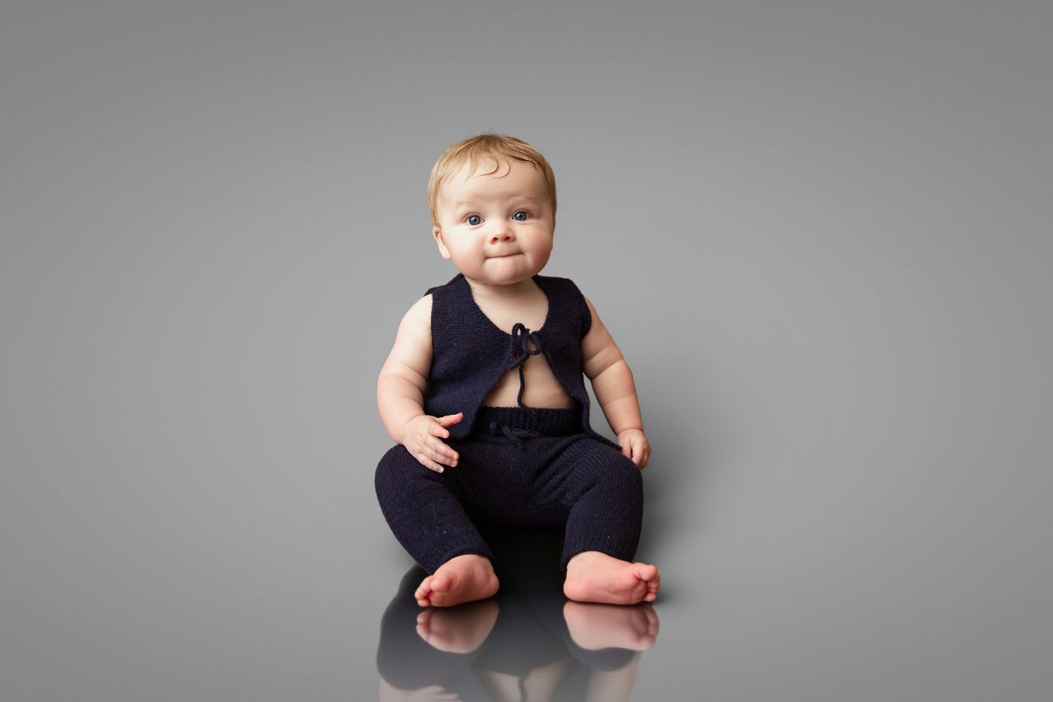 Adorable baby in navy blue romper sitting on gray studio floor, looking at camera with curious expression.