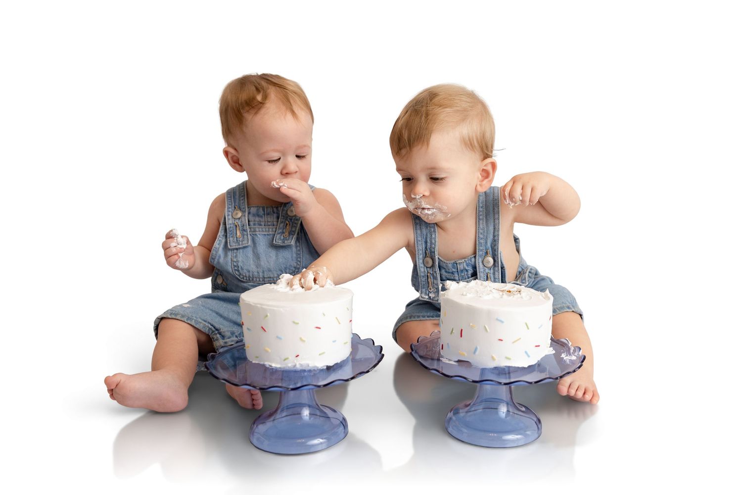 Two toddlers in denim overalls sit on a white background, each eating a small white smash cake on a blue pedestal cake stand.
