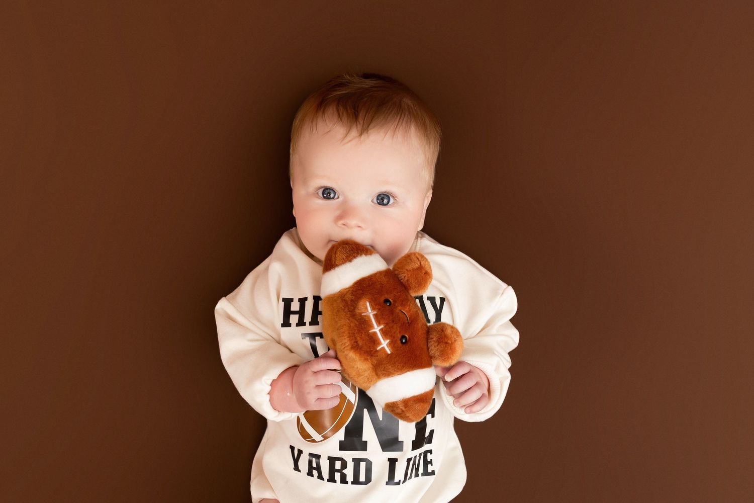 Baby wearing a white onesie with text holds and bites into a sandwich against a brown background.