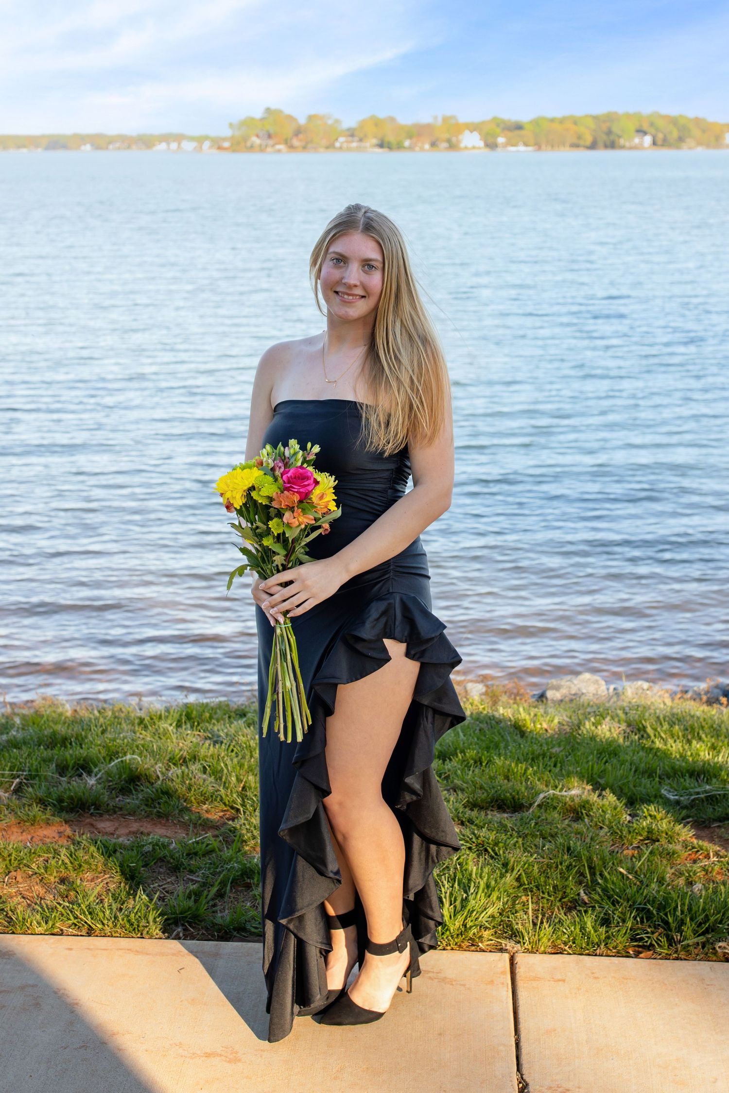 Young woman in black strapless ruffle dress holding colorful bouquet, posing by a scenic lakeside on a sunny day.