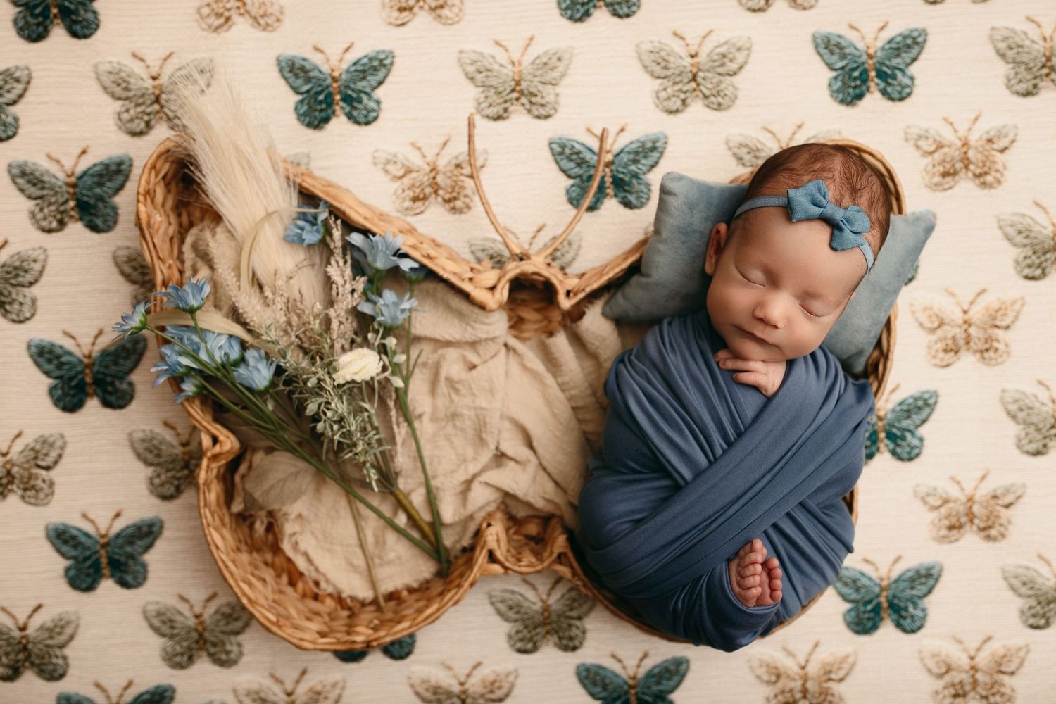 Newborn baby swaddled in blue wrap with matching headband sleeping in a woven basket beside dried flowers on a butterfly patterned backdrop.