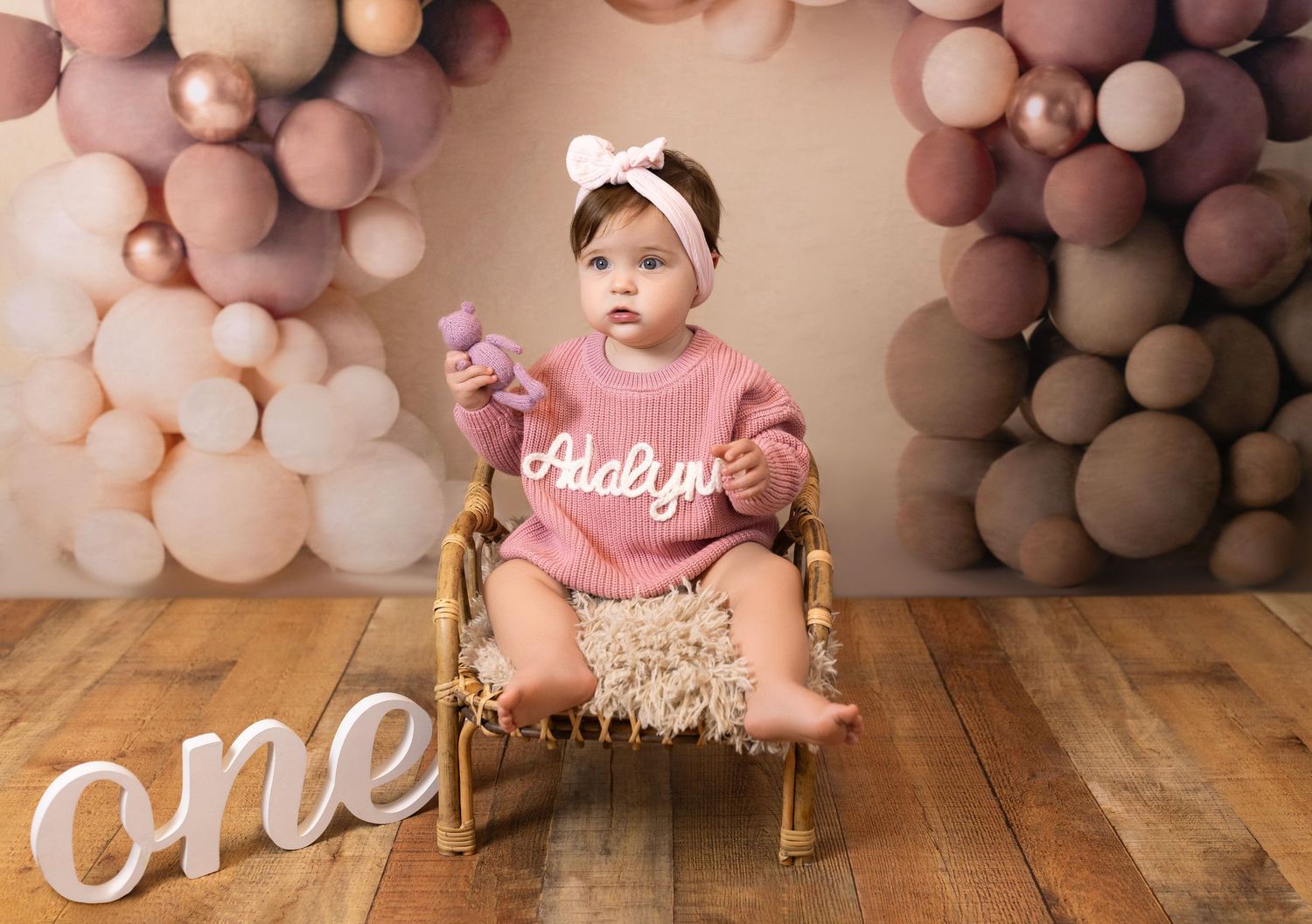 Baby in pink dress sits on wooden floor with balloon arch backdrop during first birthday photoshoot.
