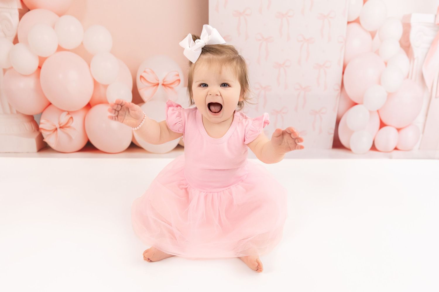 Baby in pink dress sits among soft pink balloons during a birthday or celebration photoshoot.