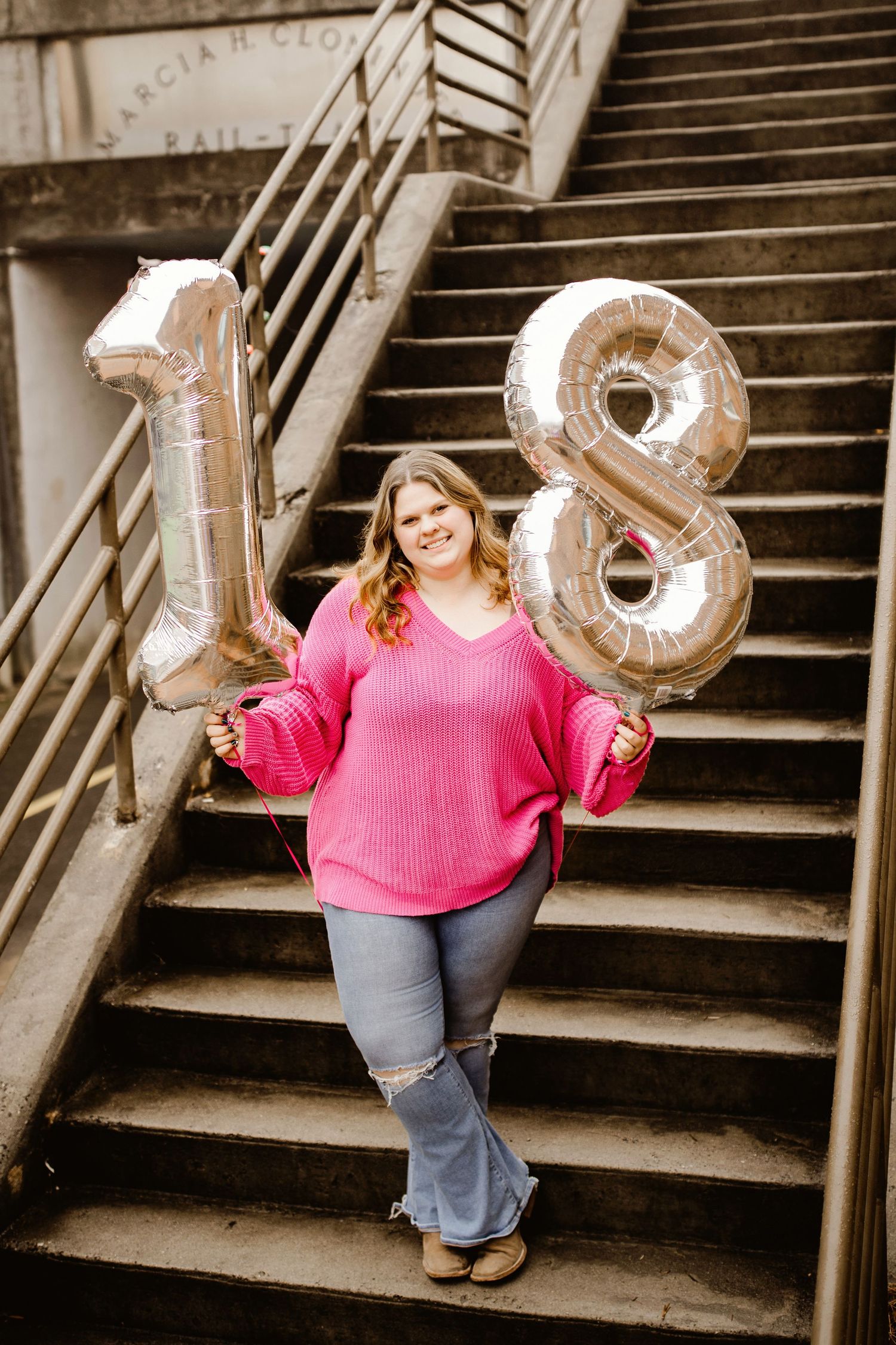 A celebratory scene on stone steps with silver number 18 balloons and someone wearing a bright pink sweater.