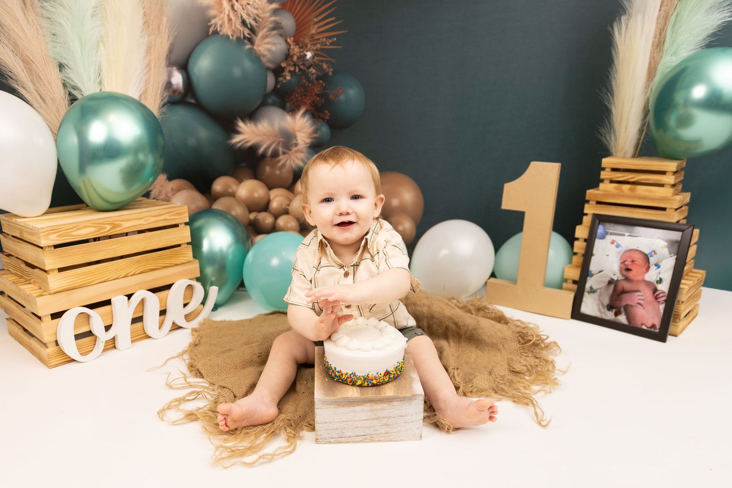 First birthday cake smash photo setup with teal and white balloons, wooden crates, and rustic decor on beige backdrop.