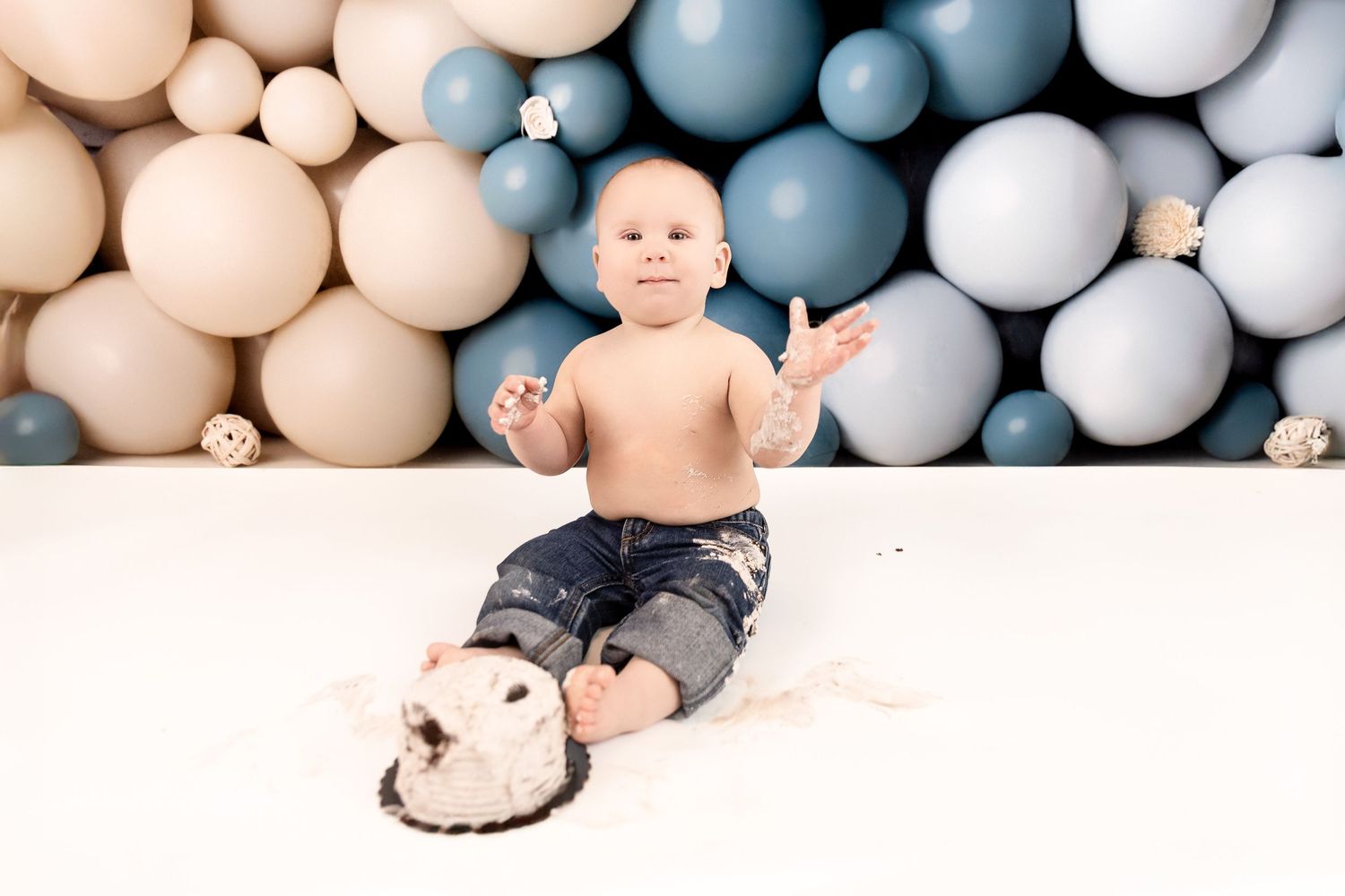 A baby lies on a white blanket surrounded by pastel blue and beige balloon decorations for a photo session.
