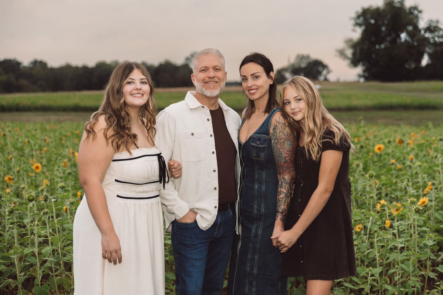 Family of four posing together in a blooming sunflower field at sunset, with a gray-haired man and three women in casual summer outfits.