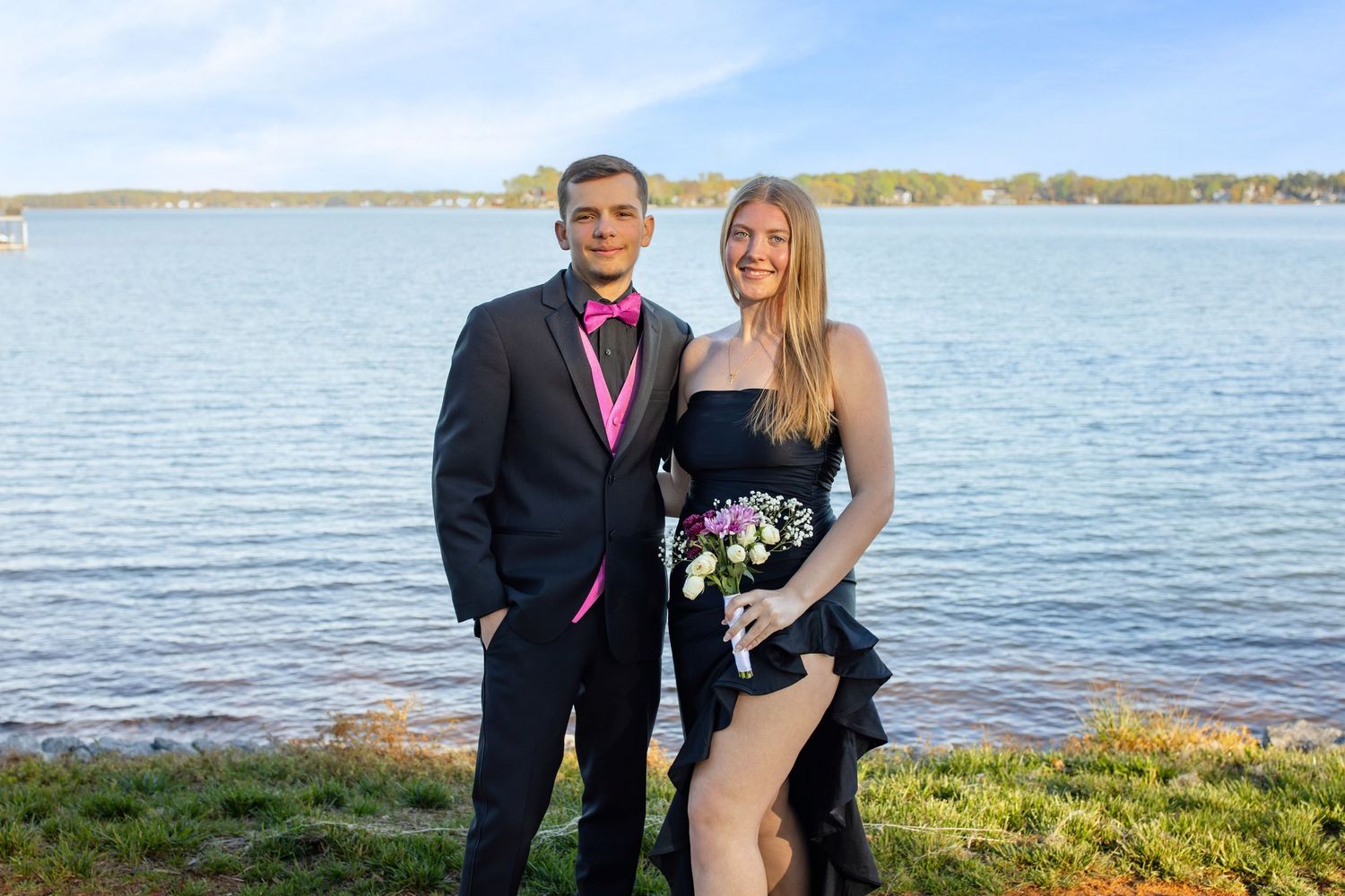 A couple dressed in prom attire poses by a lake, man in black tuxedo with pink tie, woman in black dress holding flowers.