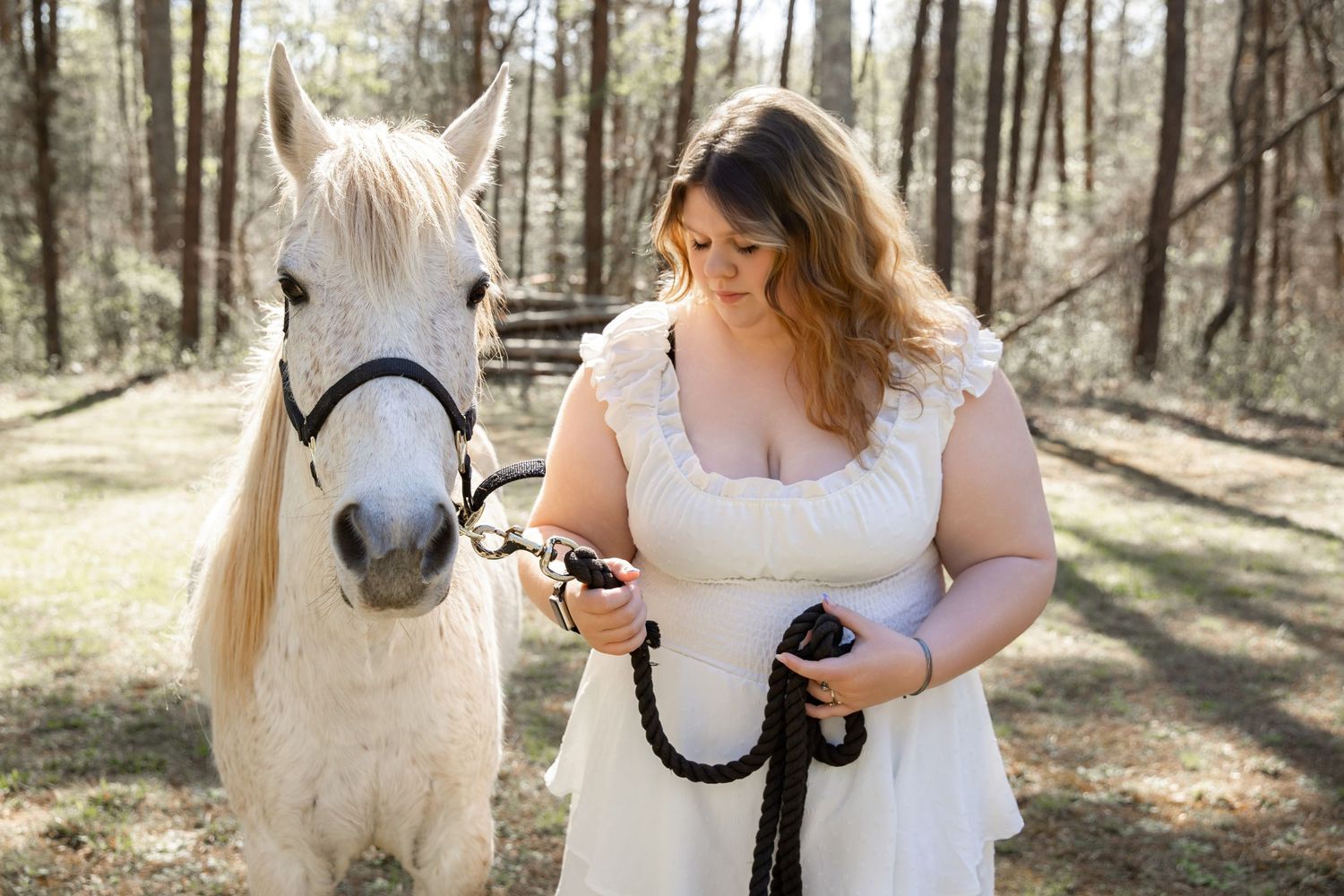 Woman in white dress holding reins of a white horse in a sunlit forest setting.