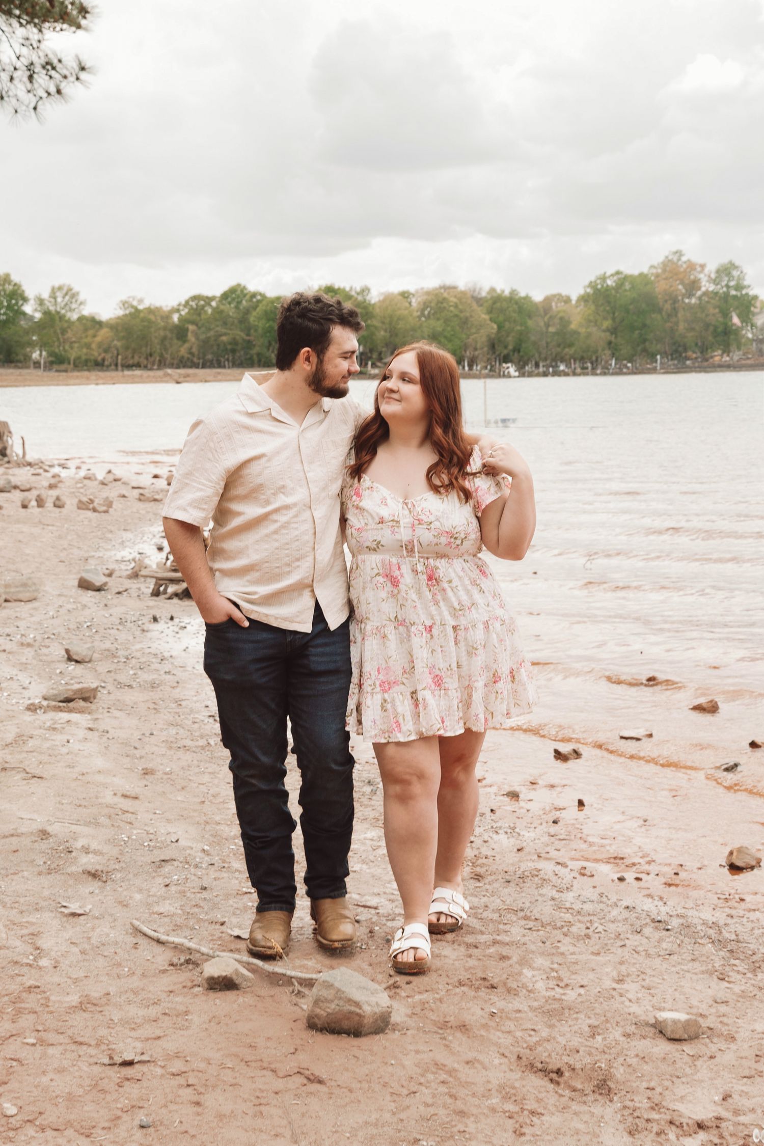 Couple walking along rocky shoreline, man in beige shirt and jeans, woman in floral dress, trees and lake in background.