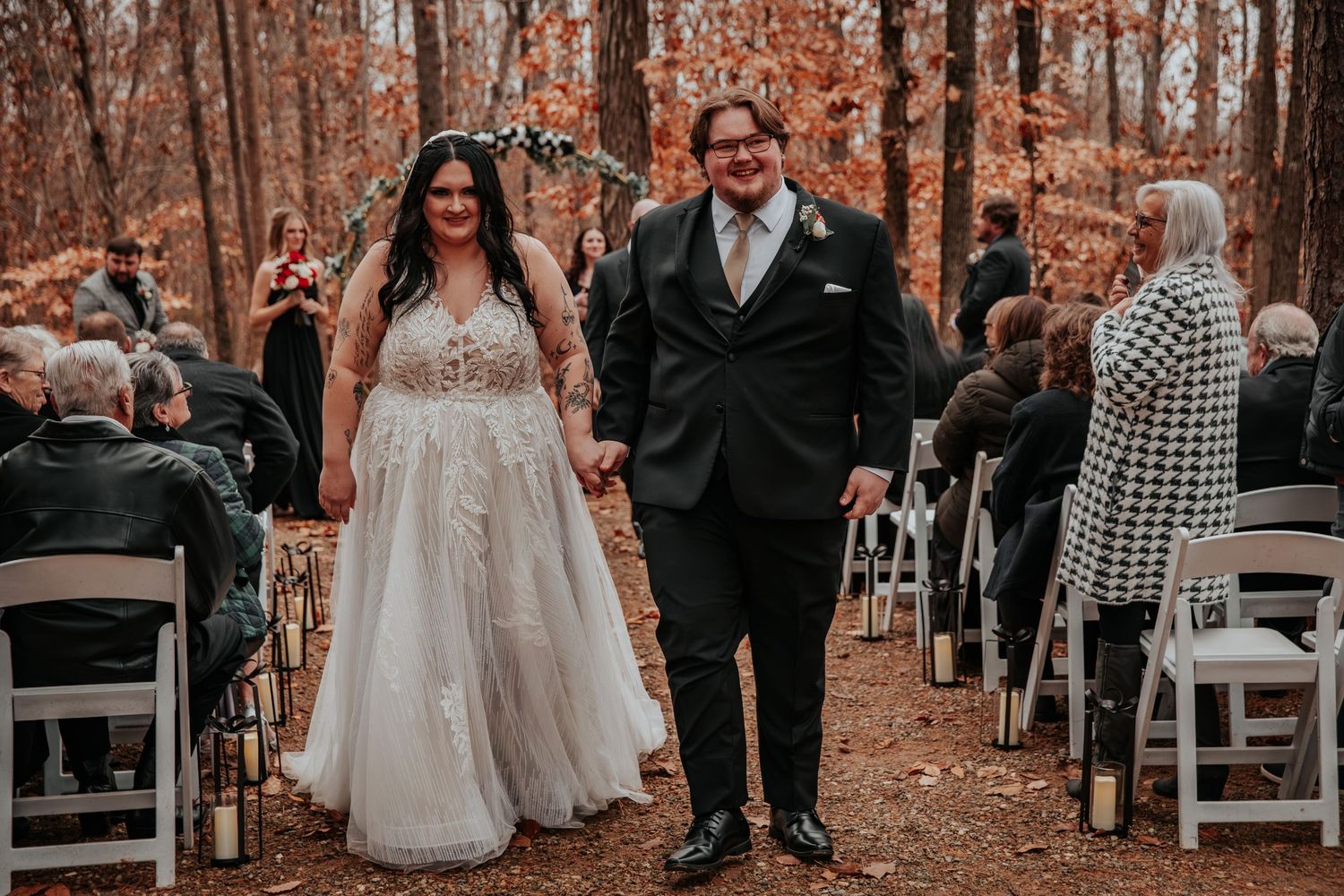 Bride and groom walk down aisle hand in hand after outdoor fall wedding ceremony in wooded setting.