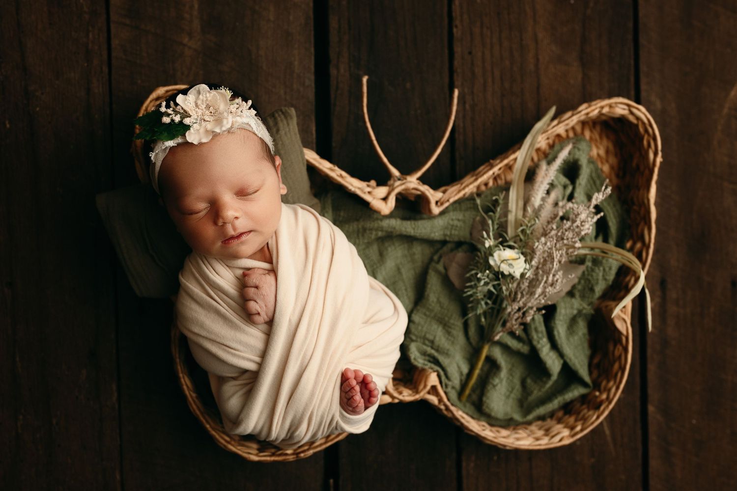 Sleeping newborn baby in a cream wrap and floral headband lying in a butterfly-shaped basket with greenery on a rustic dark wood background.