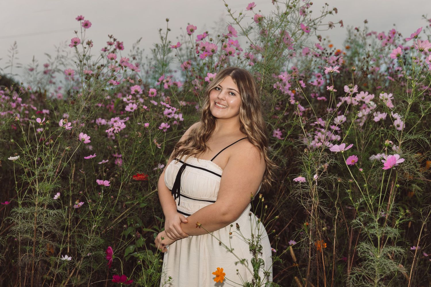 Smiling woman with long wavy hair in a white and black sundress poses among pink wildflowers in a blooming field at sunset.