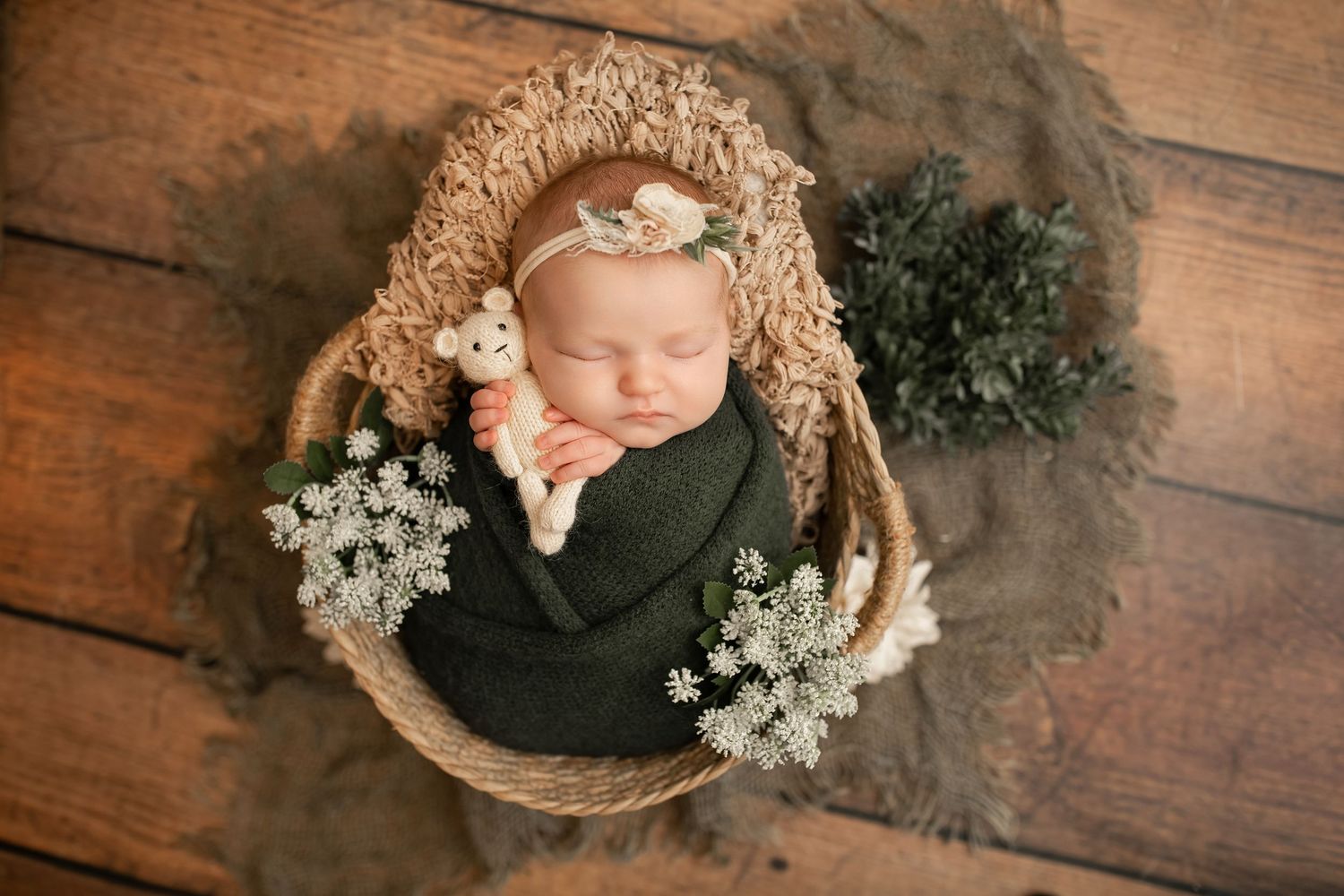 Newborn baby sleeping in wicker basket wrapped in green, holding toy, adorned with white flowers and floral headband.