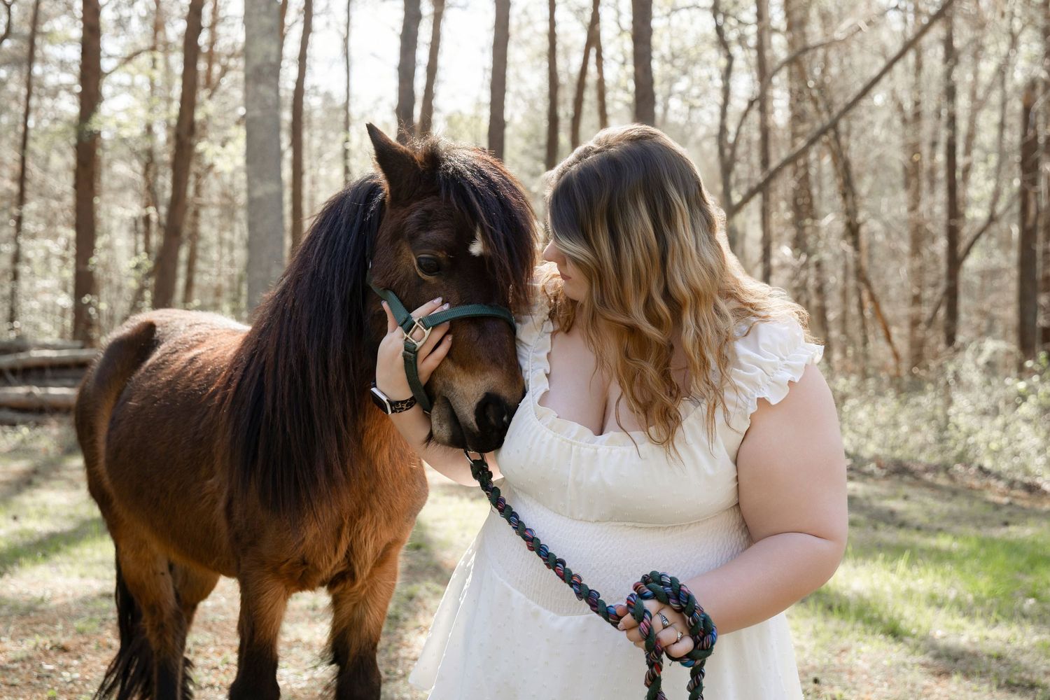 Woman in white dress bonding with a brown horse in a sunlit forest setting.