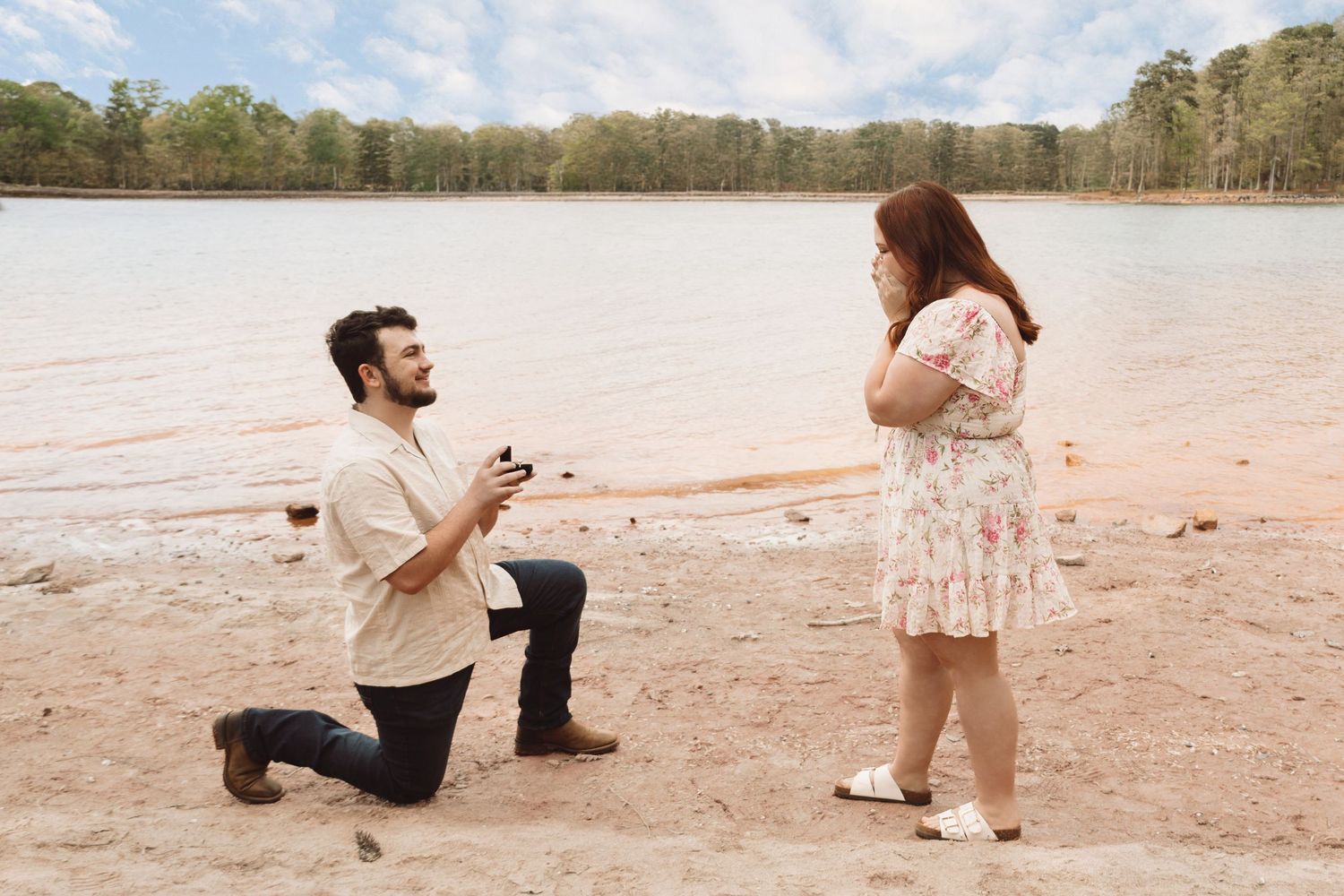 Man proposes to woman on one knee at a sandy lakeside beach surrounded by trees.