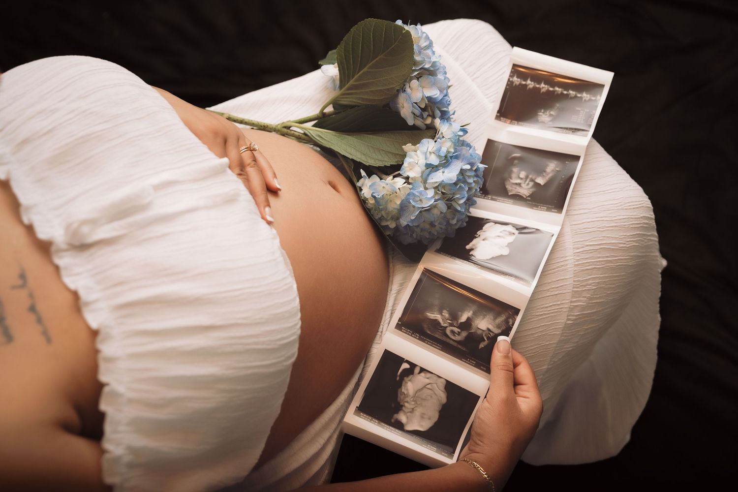 Pregnant woman in white dress holding ultrasound photos with blue hydrangeas resting on her baby bump.