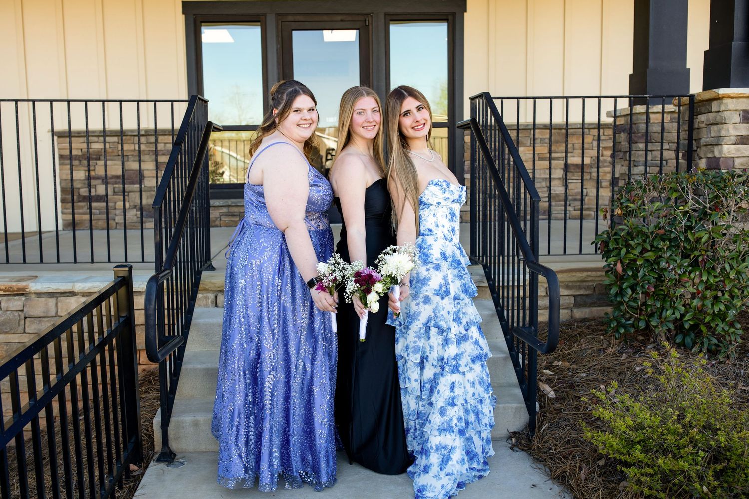 Three young women in elegant prom gowns pose together on steps, wearing blue and black formal dresses with bouquets.