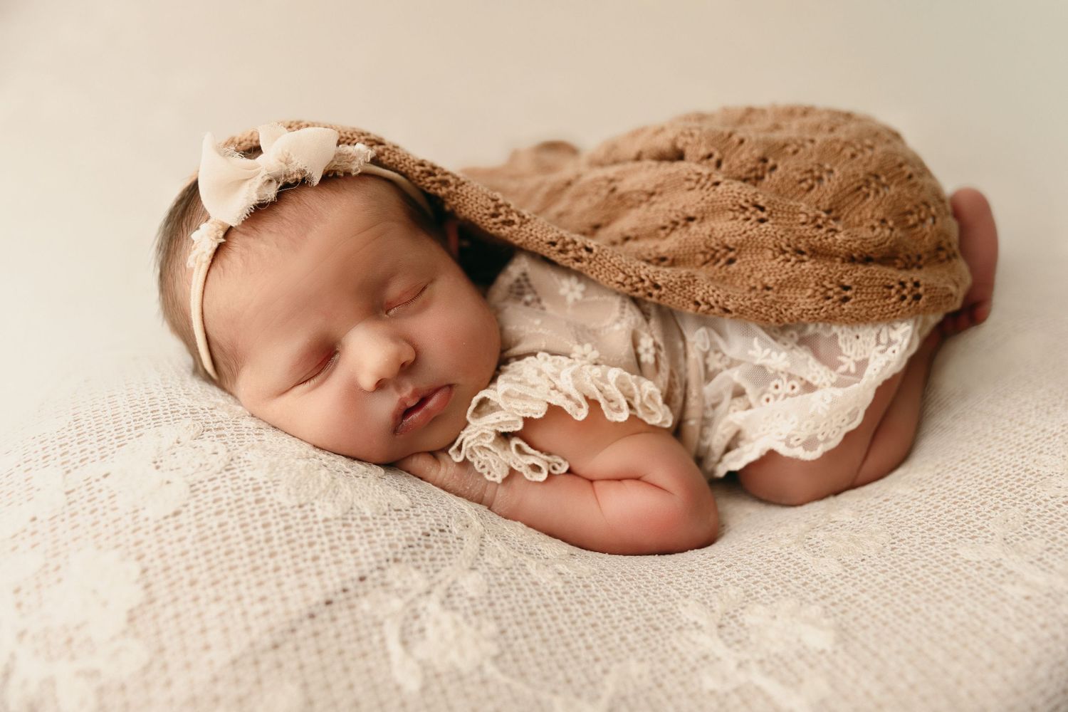Sleeping newborn baby in a lace outfit, bow headband and brown knit wrap, posed on a cream lace blanket in a newborn photo session.