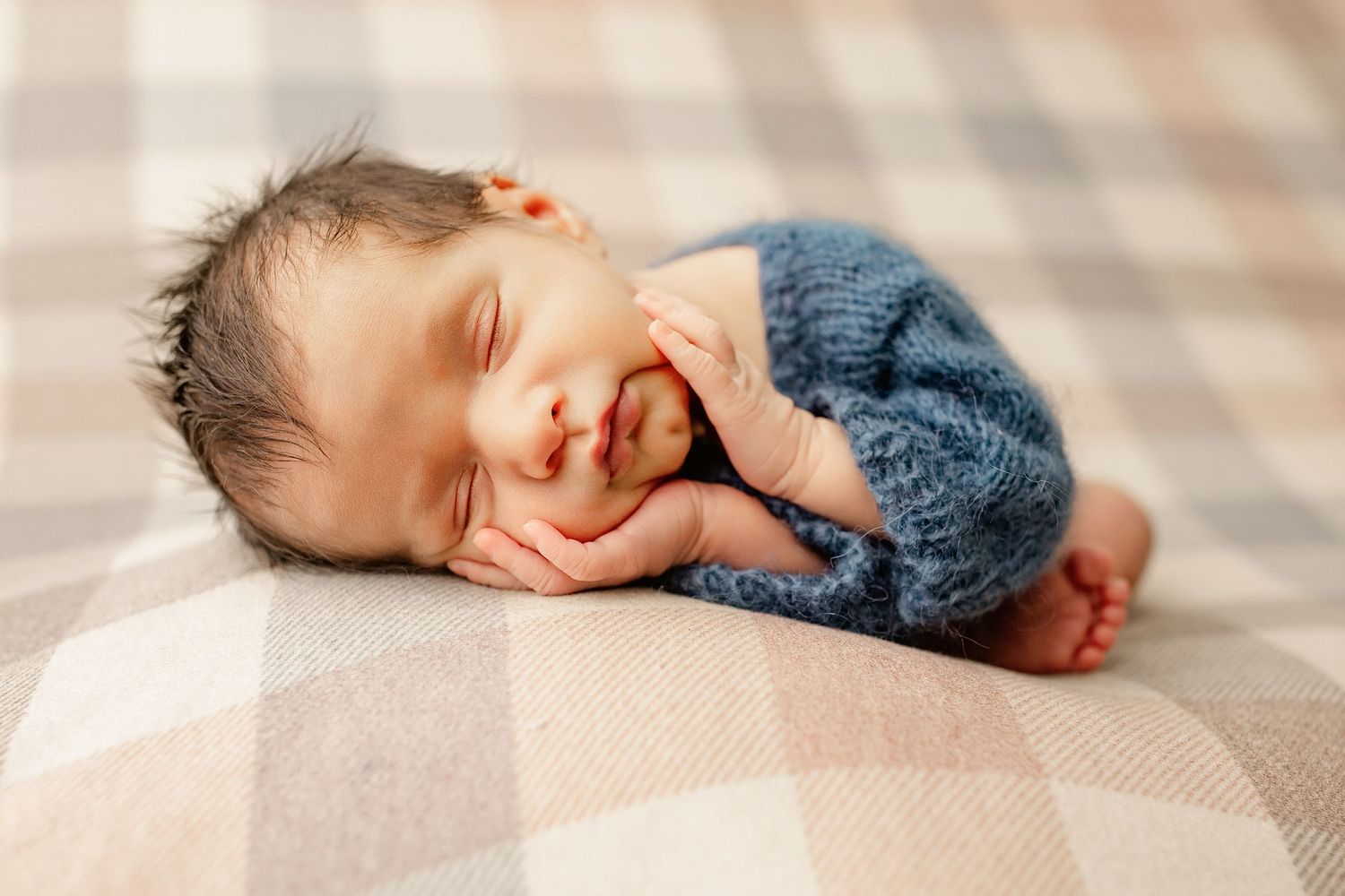 A newborn baby wearing a blue knit romper sleeps peacefully on a plaid blanket.