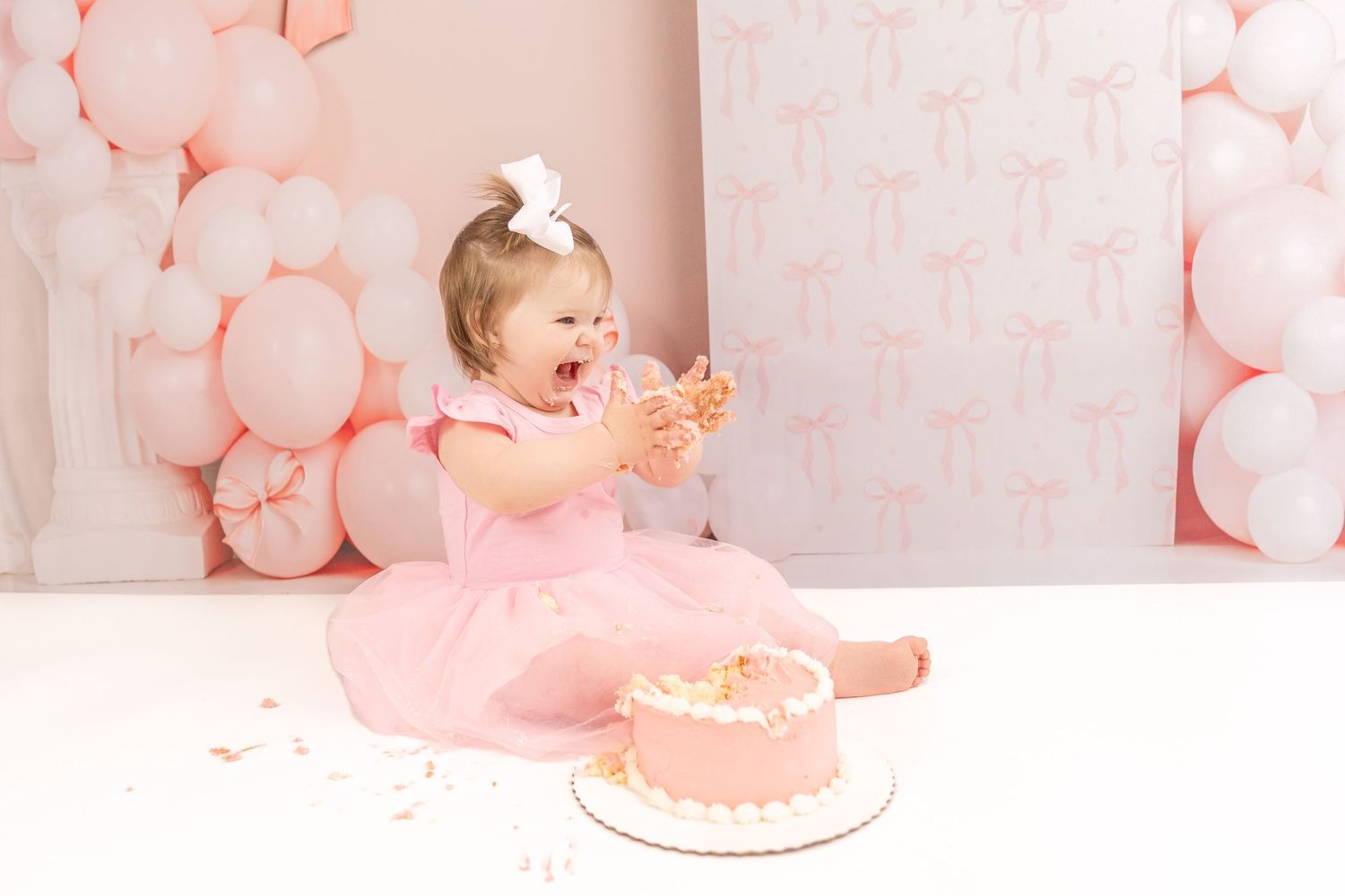 Pink balloon garland decorates a first birthday party setup with cake and floral accents in soft pastel colors.