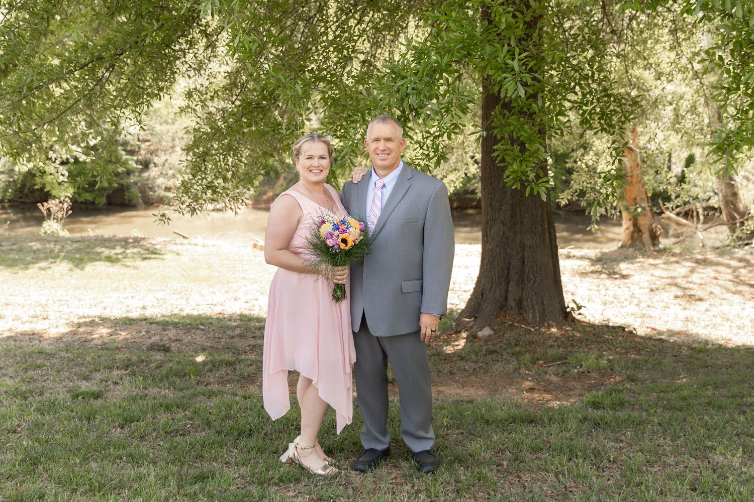 Couple posing outdoors under a large tree, woman in pink dress holding bouquet, man in gray suit, at a wedding.