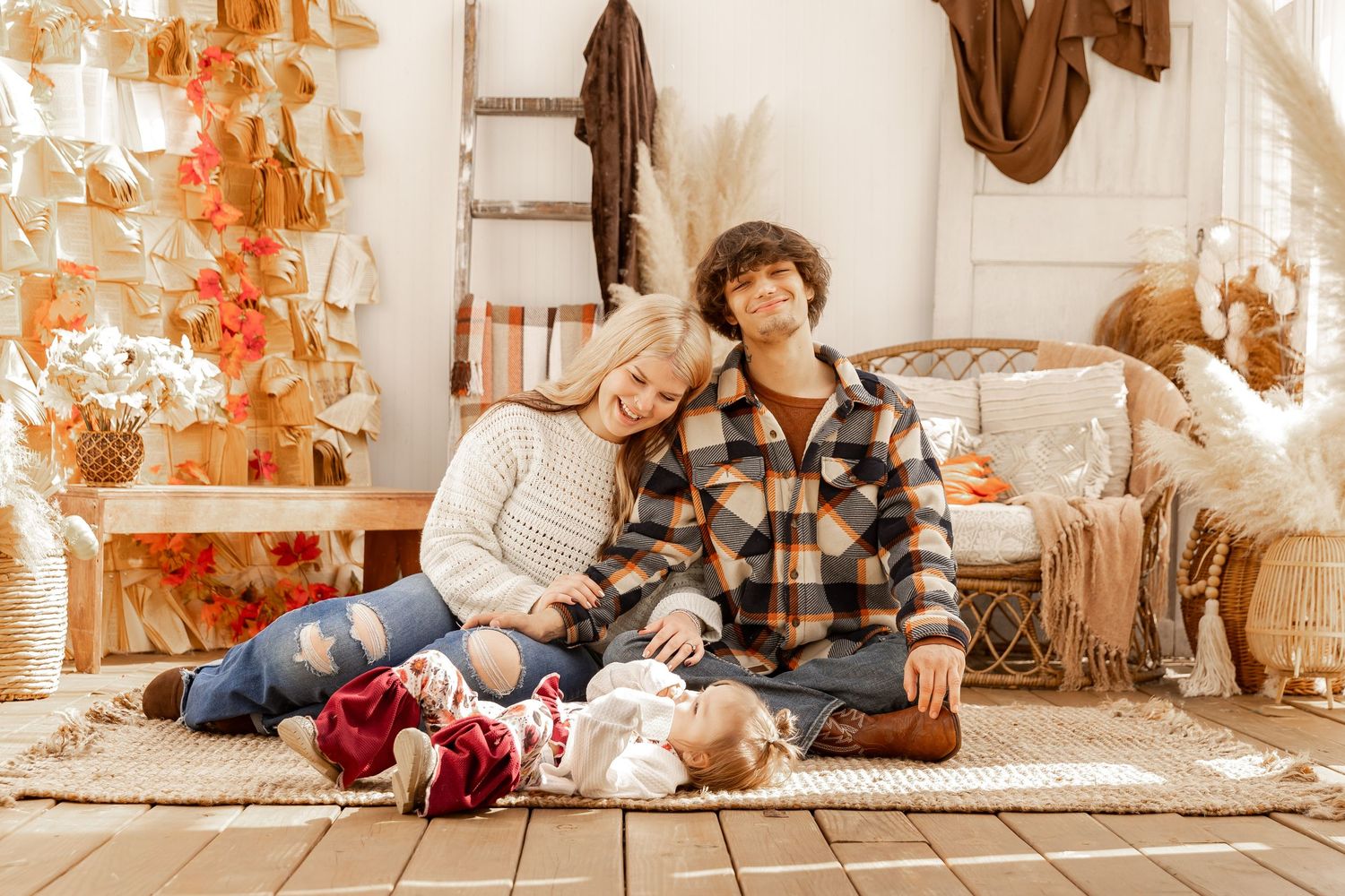 Smiling couple in cozy flannel and sweater sit on a boho rug with their baby during a warm autumn family photo session.