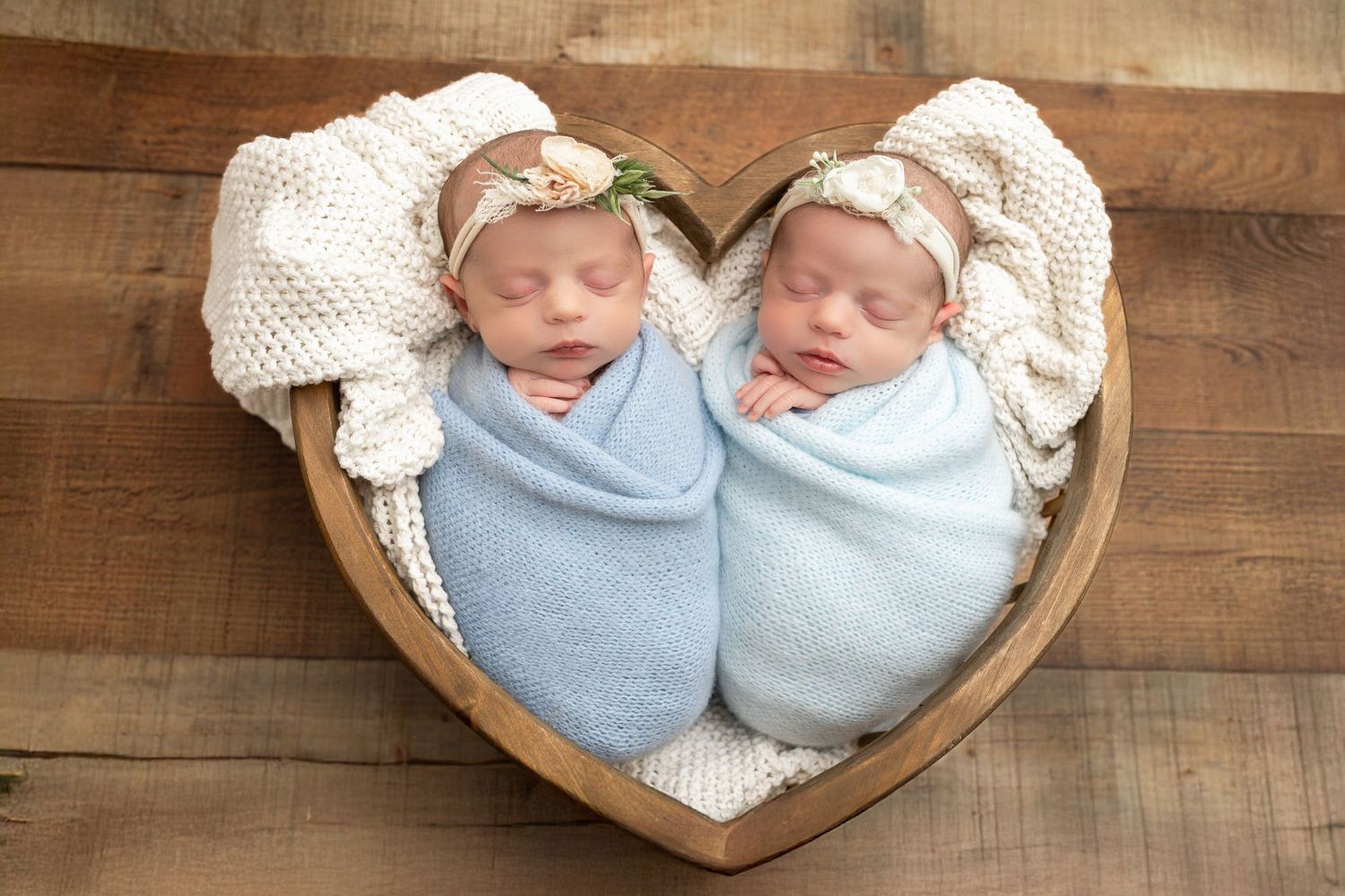 Two sleeping newborn babies swaddled in blue blankets lie together in a heart-shaped basket against wooden backdrop.
