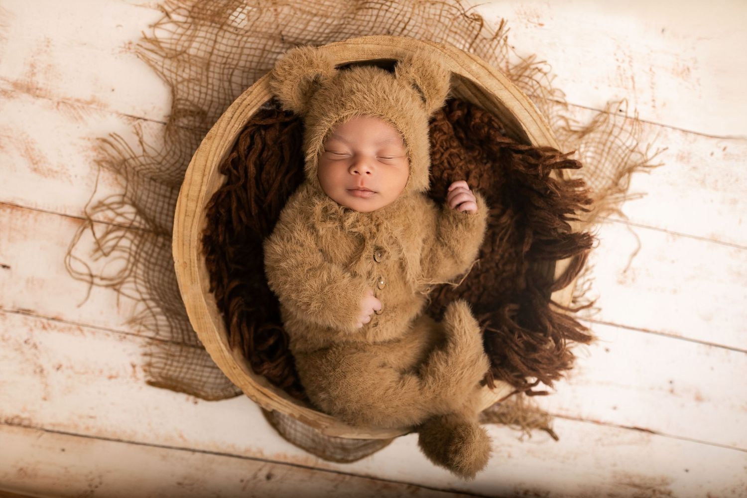 Newborn baby sleeping in a round wooden basket wearing a brown teddy bear outfit and matching hat.
