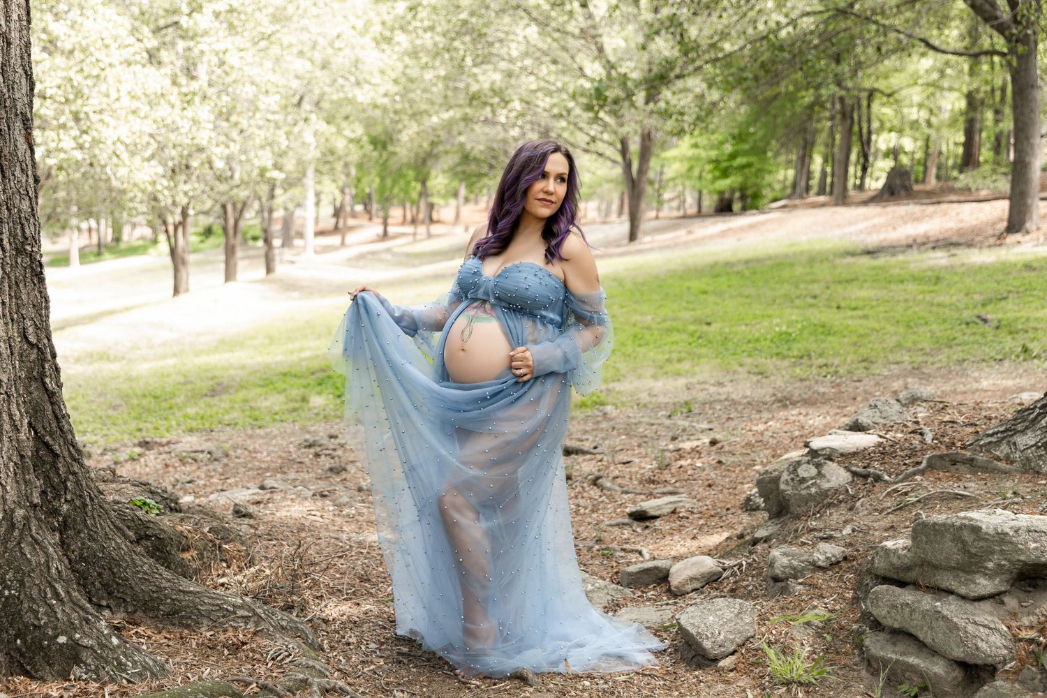Pregnant woman in blue tulle maternity gown posing outdoors in a sunlit park with trees and stone wall.