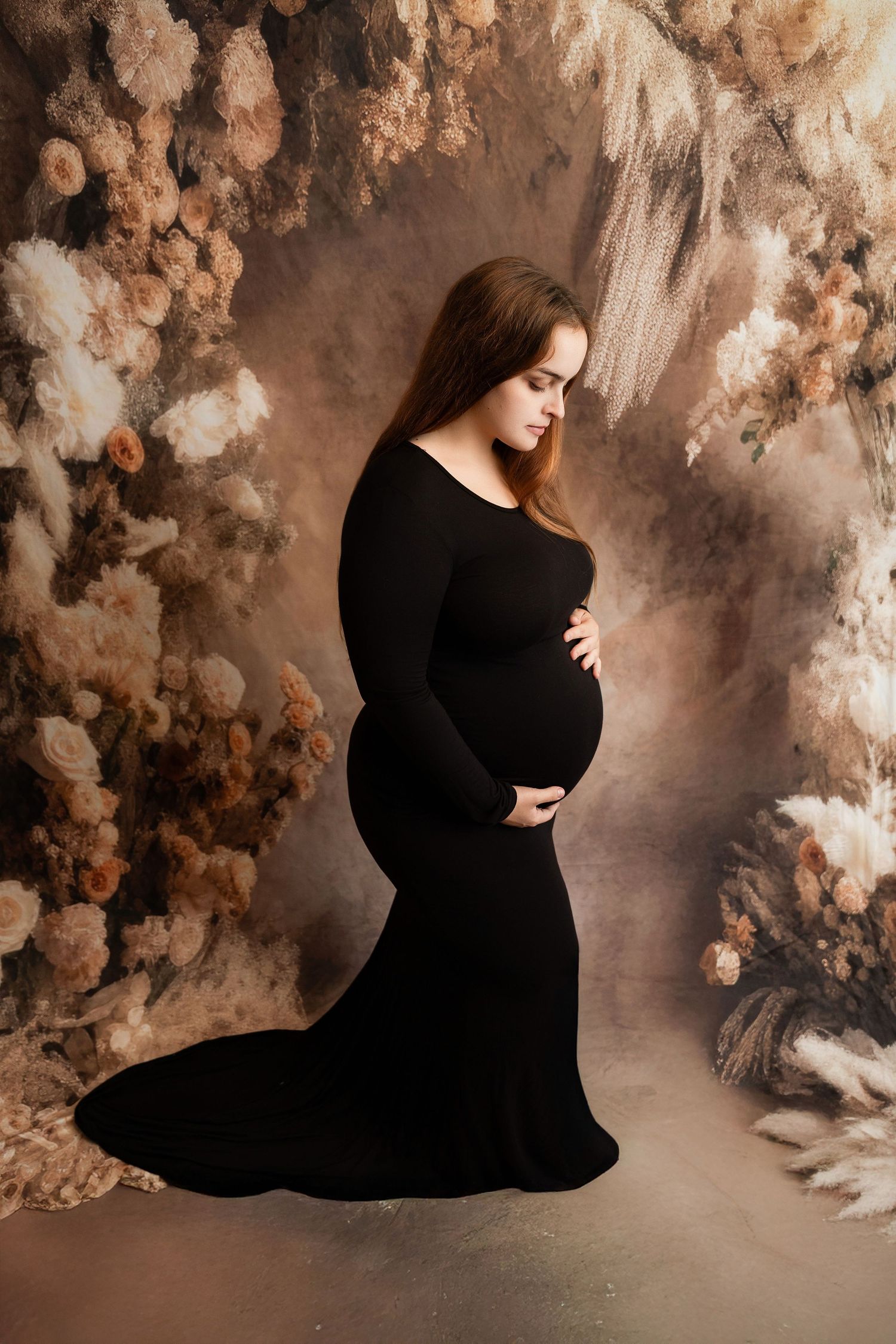 Pregnant woman in elegant black gown cradling baby bump in front of floral backdrop.