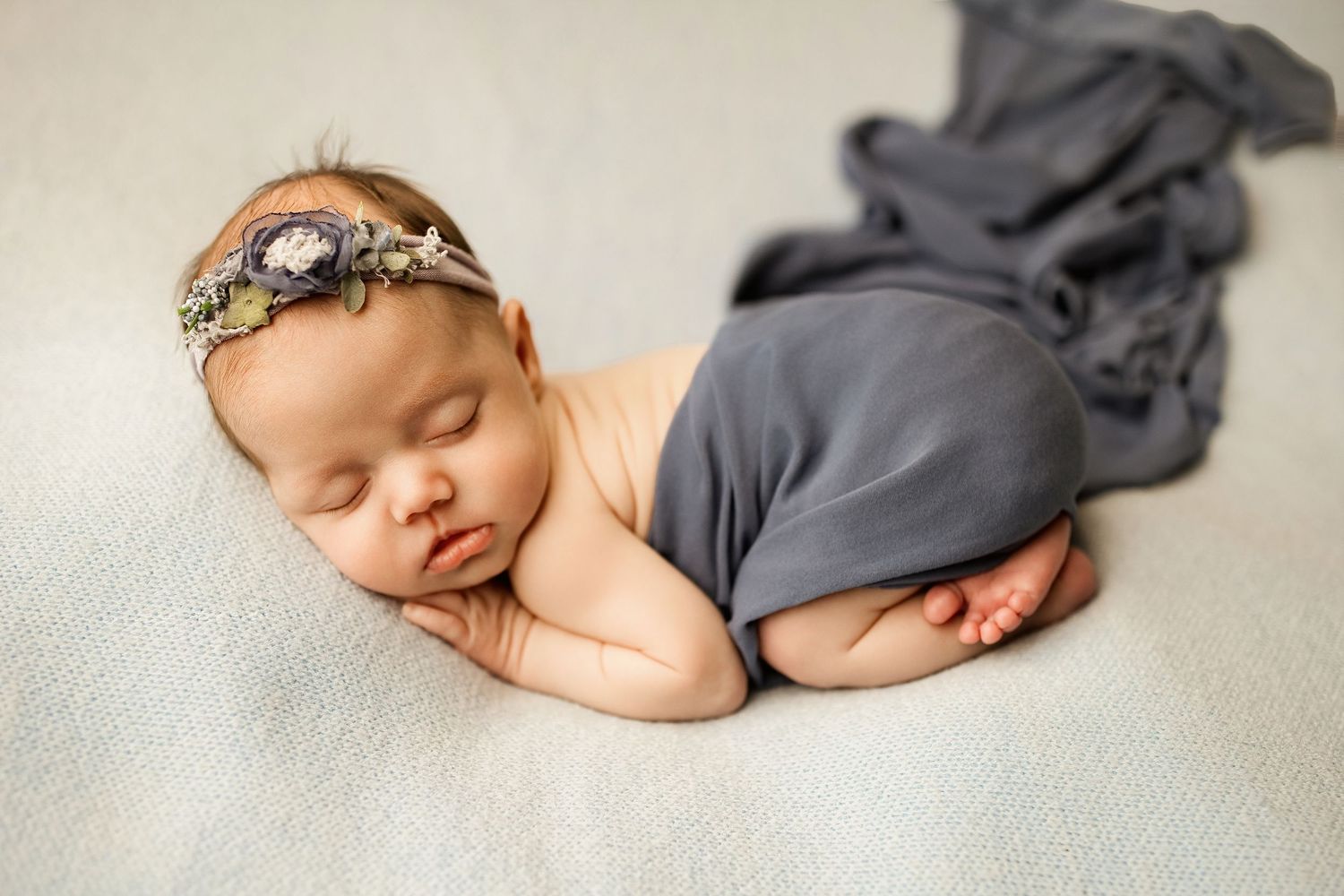 A sleeping newborn baby wearing a gray wrap and headband lies peacefully on a soft white blanket.