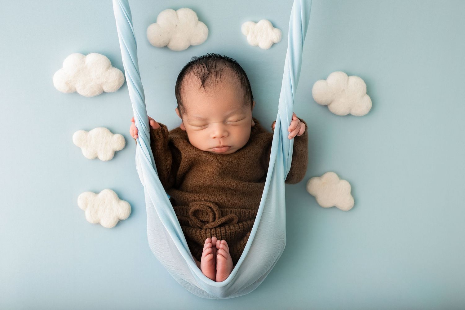 Newborn baby sleeping peacefully in a blue fabric swing, wrapped in brown, surrounded by white cloud decorations.