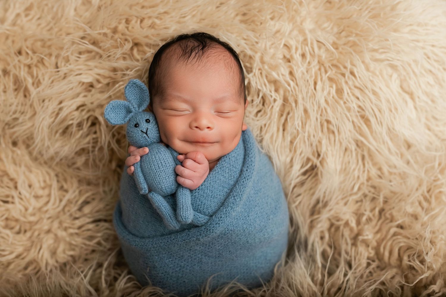 Smiling newborn baby swaddled in blue wrap holding a knit bunny toy on a fluffy beige fur backdrop.