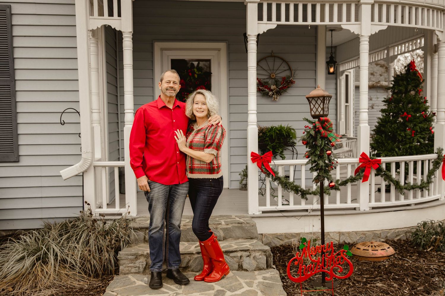 A couple in coordinating red and black outfits poses for holiday photos in front of a white house with festive decorations.